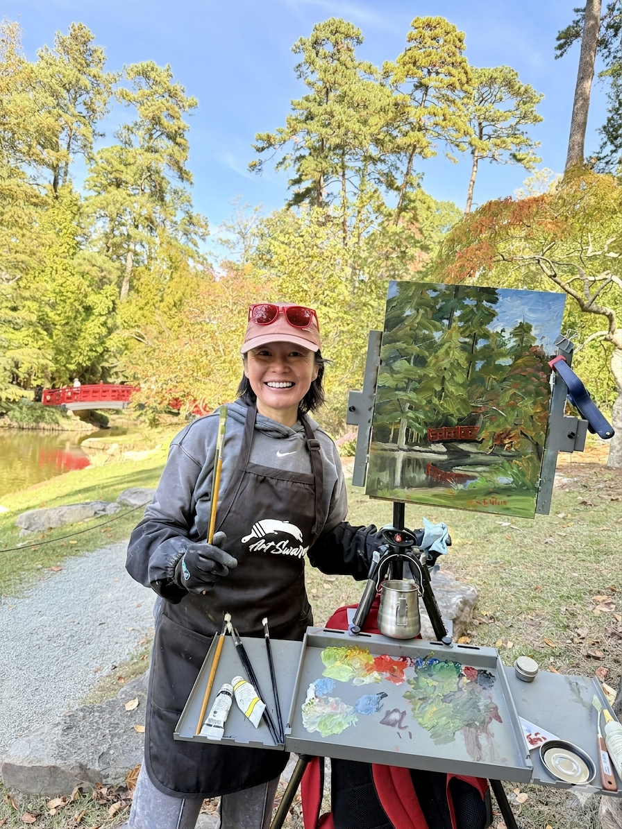 A woman artist outdoors in a park, standing at her easel, smiling, with a landscape painting of trees and a red bridge, surrounded by colorful autumn foliage.