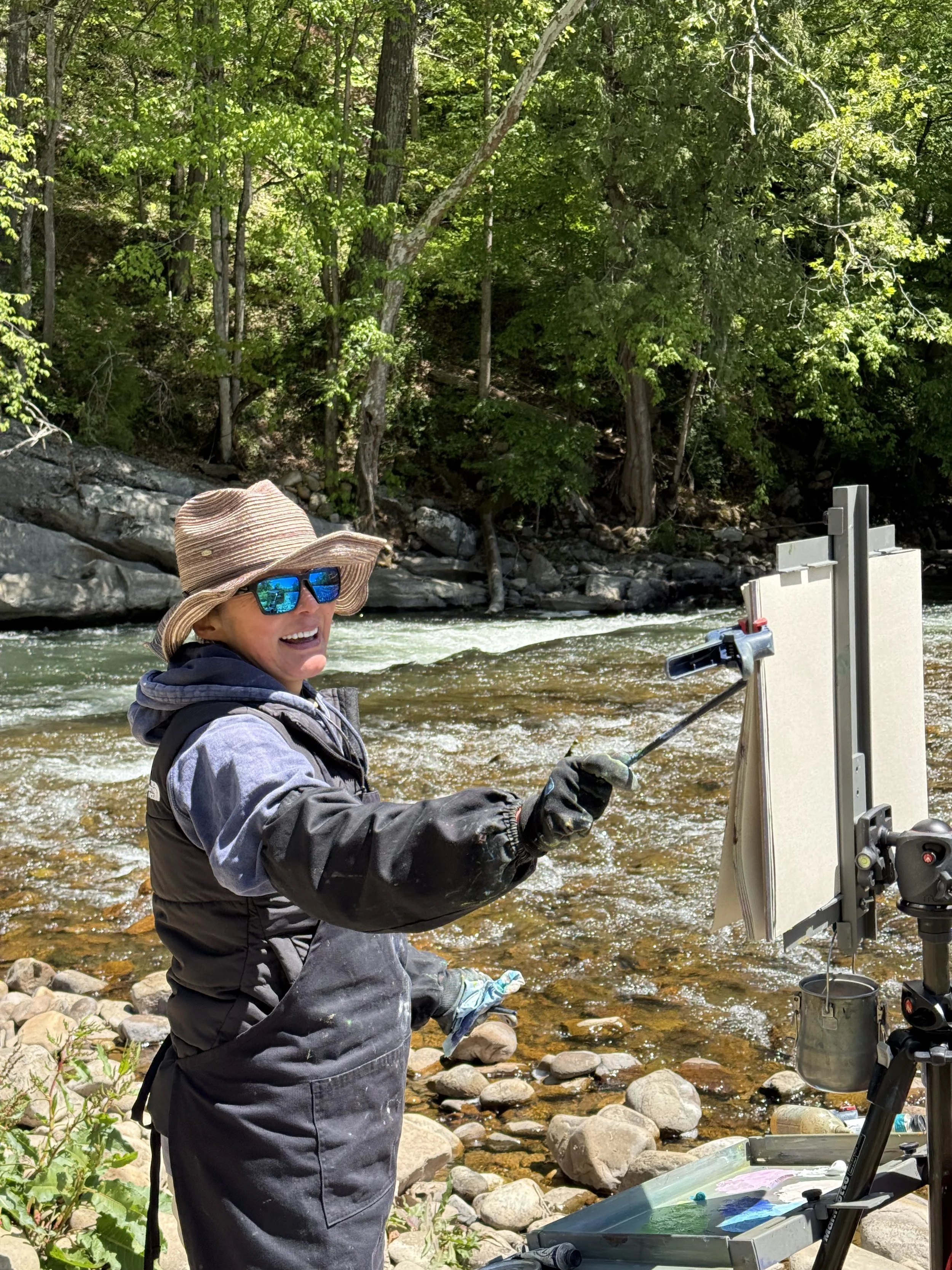 Woman working outdoors near a river wearing sunglasses, a large hat, and waterproof clothing, operating equipment on a stand, with trees and rocks in the background.