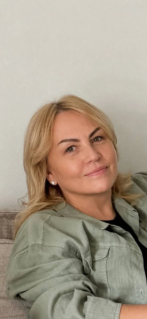 A woman with blonde hair and earrings sitting on a couch against a plain wall.