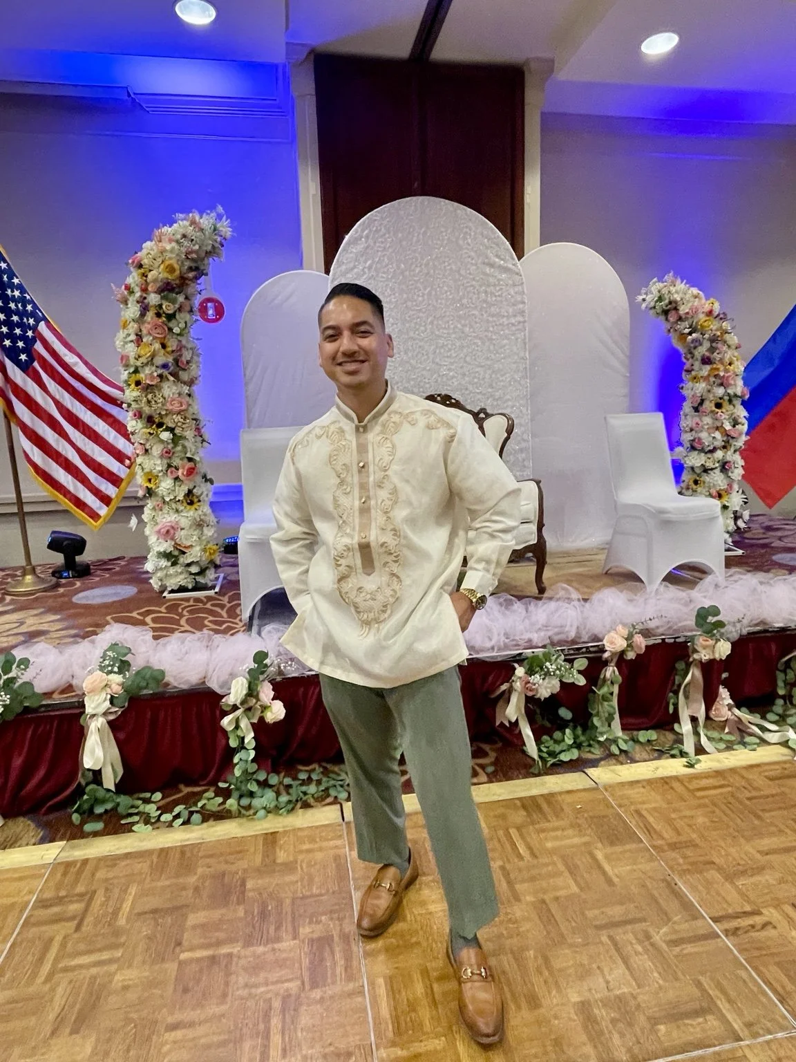 A man wearing a beige traditional shirt with embroidery, khaki pants, and brown loafers stands in front of a decorated stage with floral arrangements and flags of the United States and Russia.