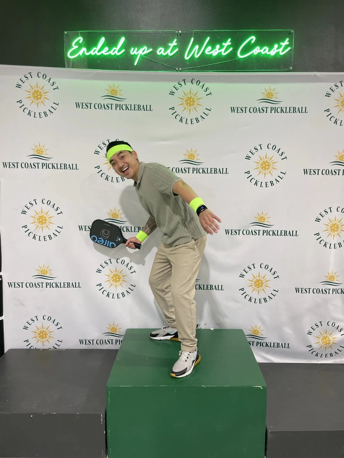 A man with a neon yellow headband and wristbands, holding a pickleball paddle, standing on a green podium at a West Coast Pickleball event, smiling and posing cheerfully.