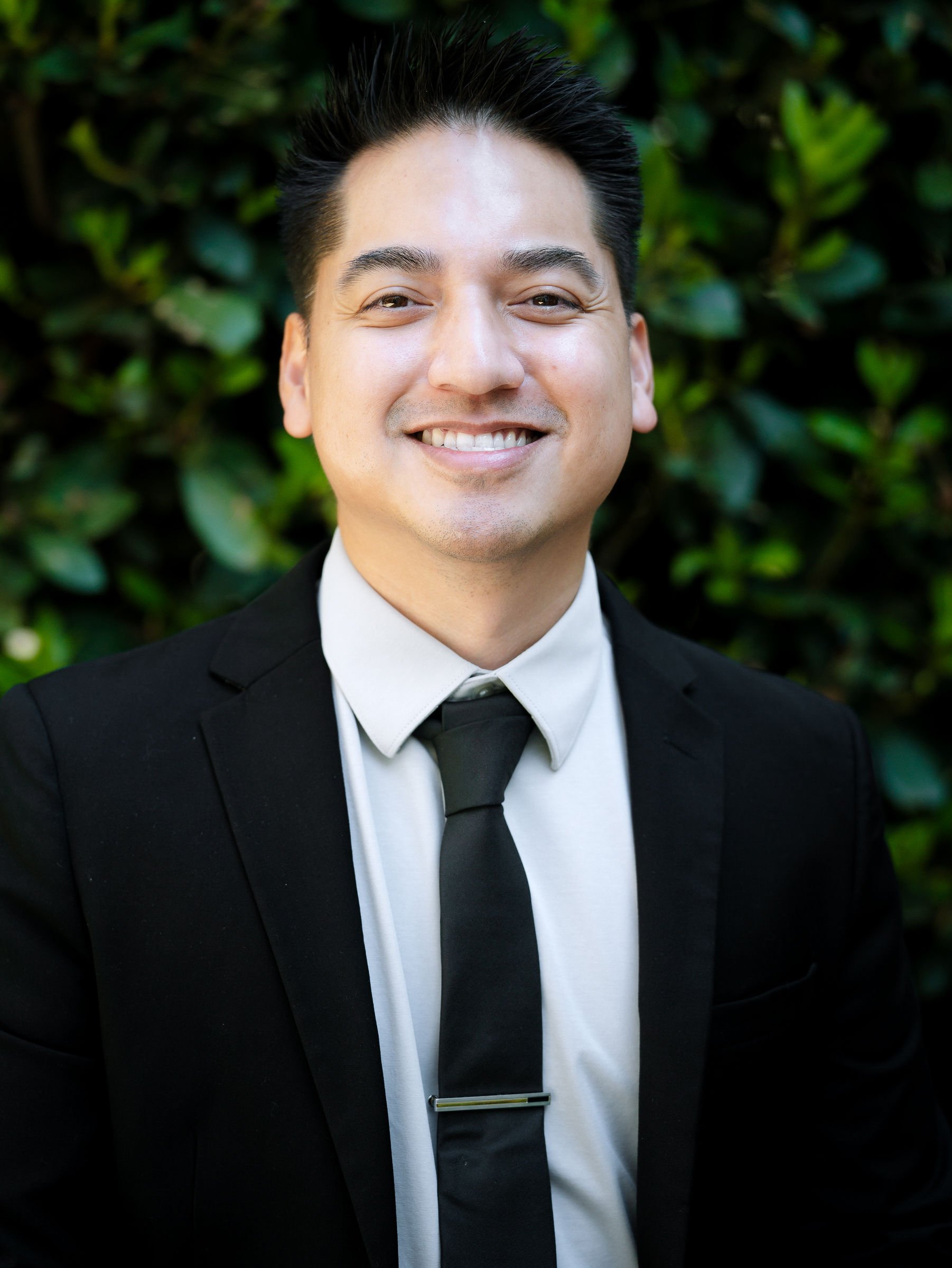 A smiling man in a tuxedo standing outdoors with blurred green foliage in the background.