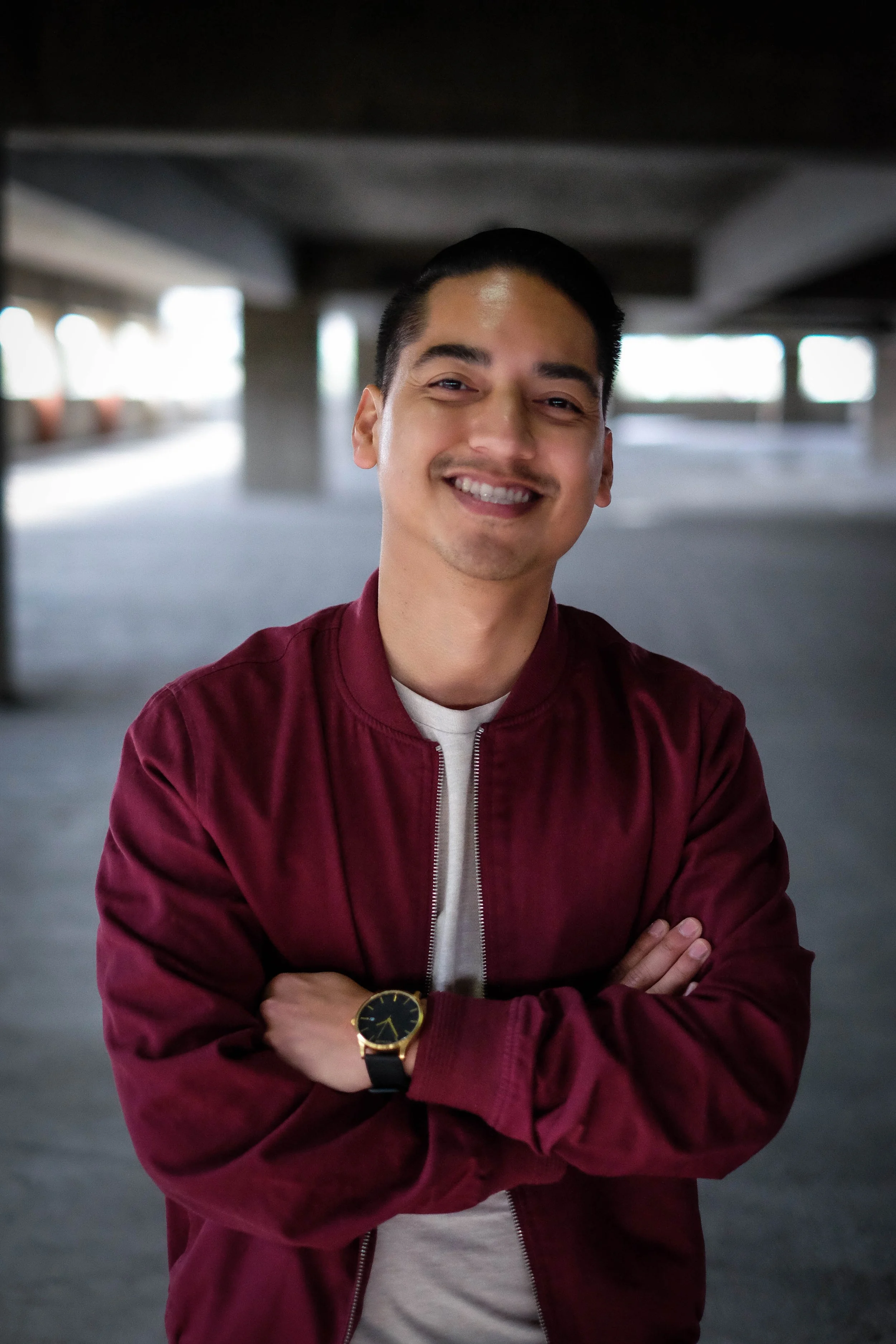 A young man with dark hair, smiling with arms crossed, wearing a burgundy jacket and a black watch, standing in an indoor parking garage.