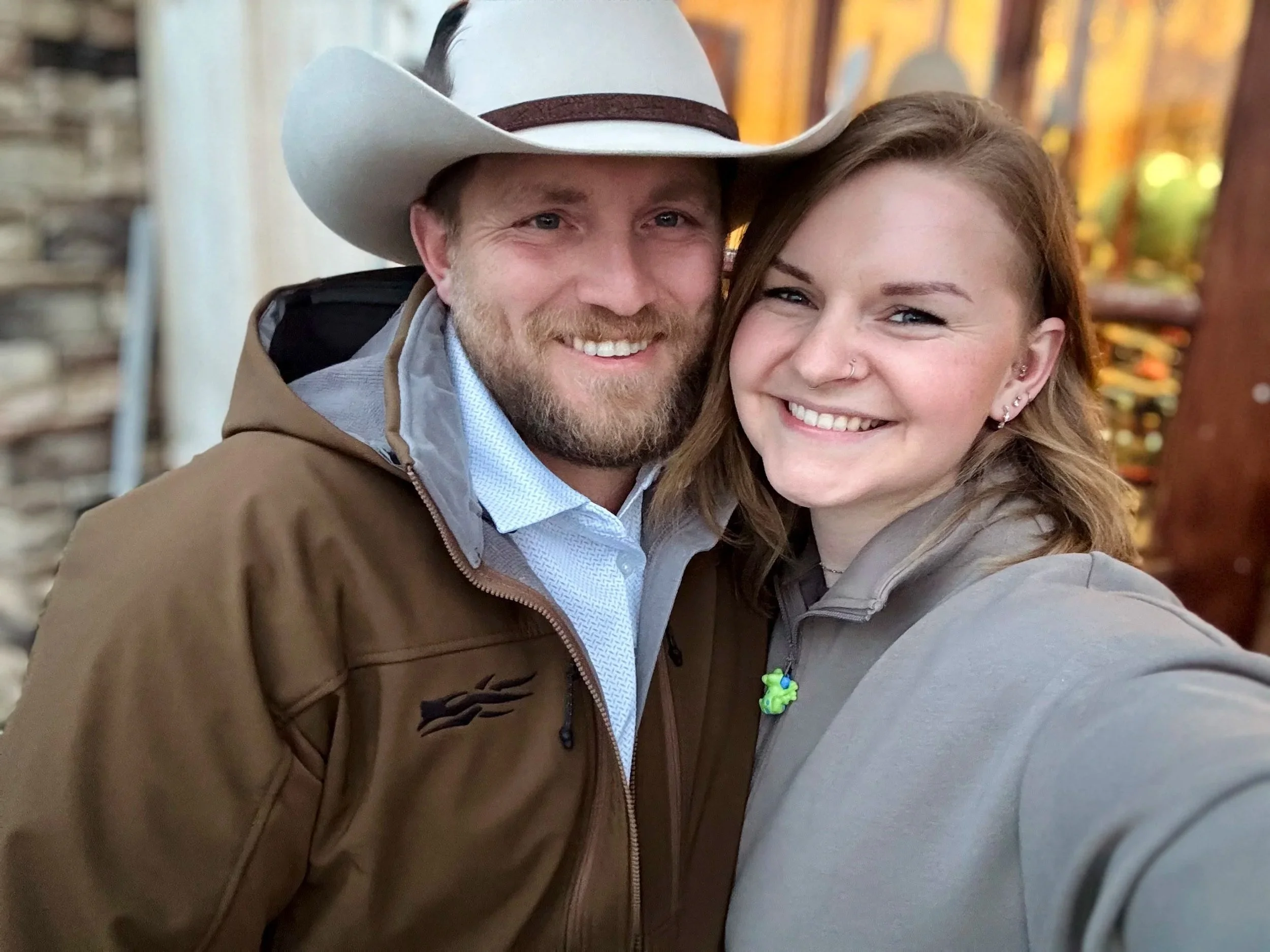 Close-up of a smiling couple taking a selfie outdoors, with trees and autumn foliage in the background. The man has a beard, wears a cowboy hat and a brown jacket. The woman has long light brown hair, a nose piercing, earrings, and is wearing a light gray jacket.