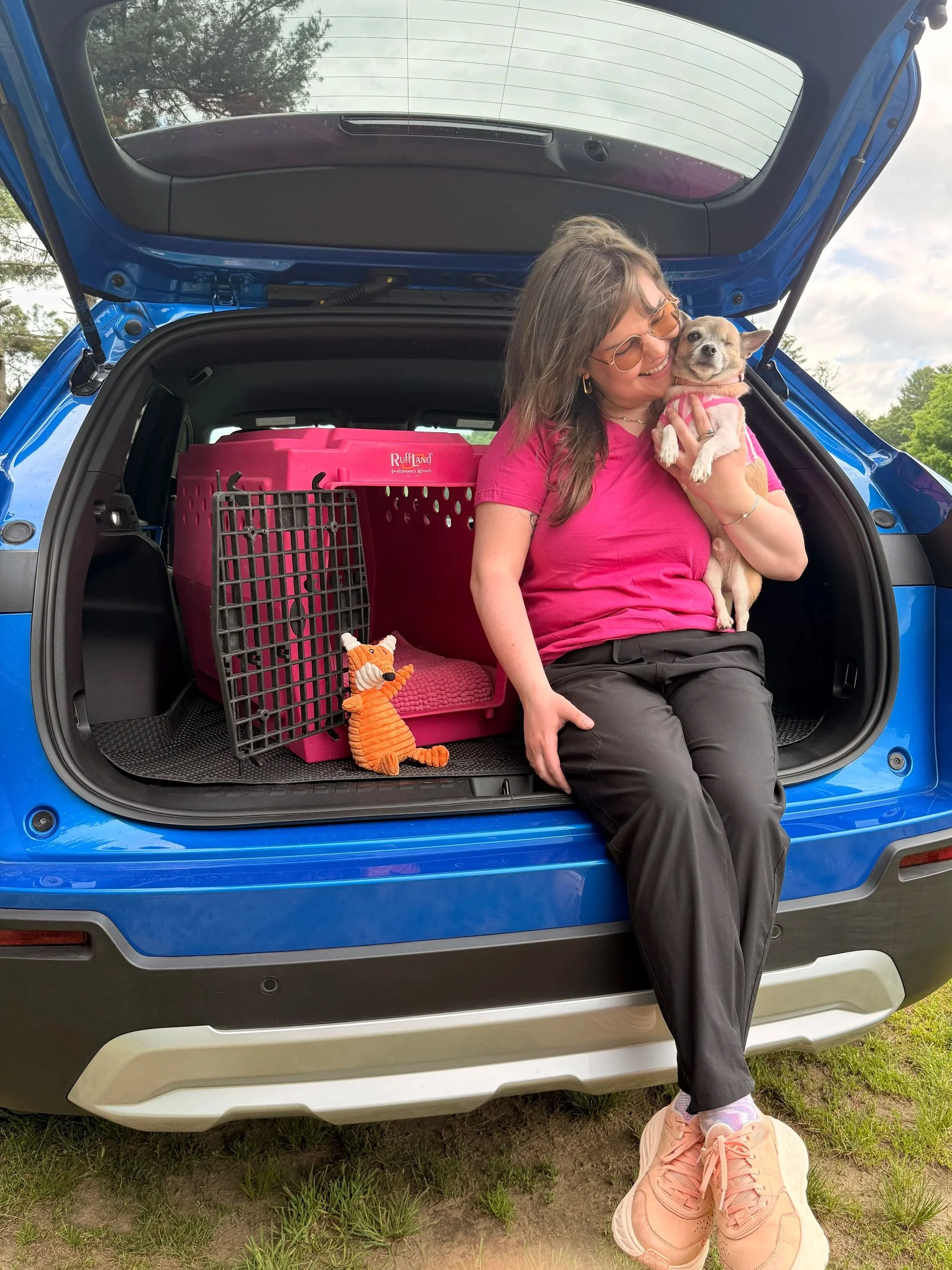 A woman is sitting in the open trunk of a blue SUV, smiling and holding a small dog. The woman is wearing glasses, a pink shirt, and black pants. There are pet carriers and toys in the trunk. The woman has pink sneakers in front of her on the ground. The background shows trees and a cloudy sky.