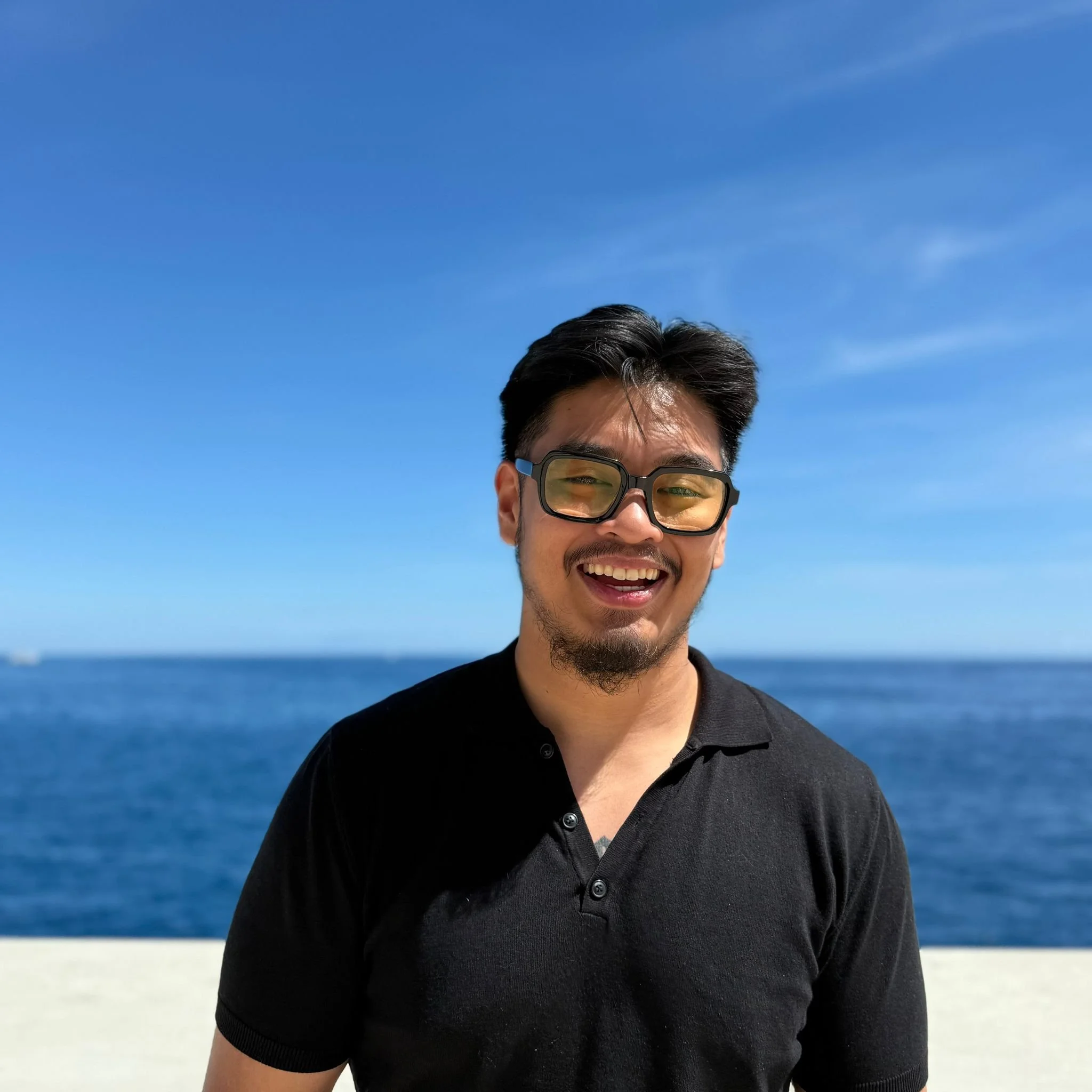 Close-up of a smiling man wearing black sunglasses and a black polo shirt, outdoors near a body of water with a clear blue sky.