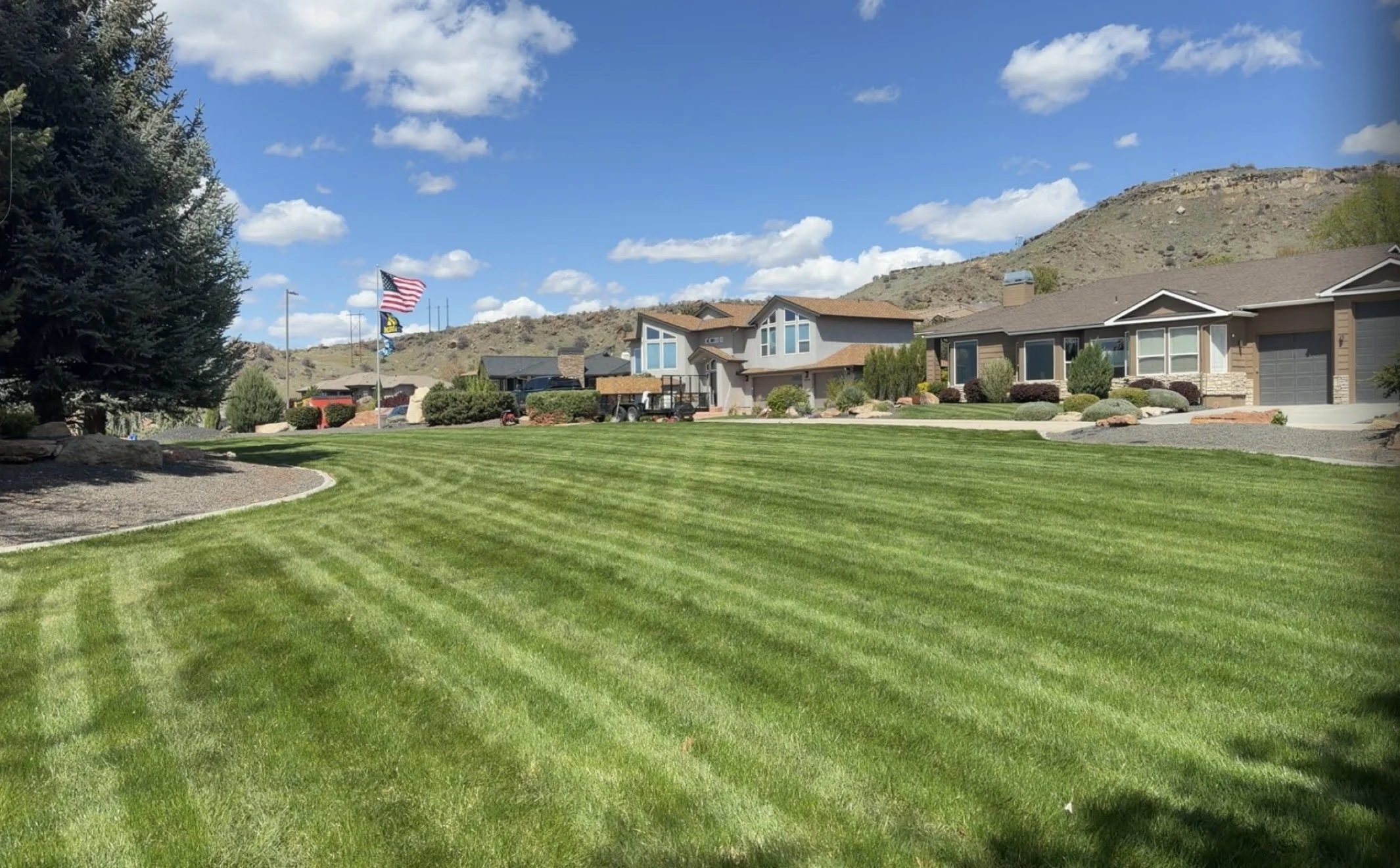 A well-maintained suburban neighborhood with modern houses, a large green lawn in the foreground, flags waving on flagpoles, and mountains in the background under a partly cloudy sky.