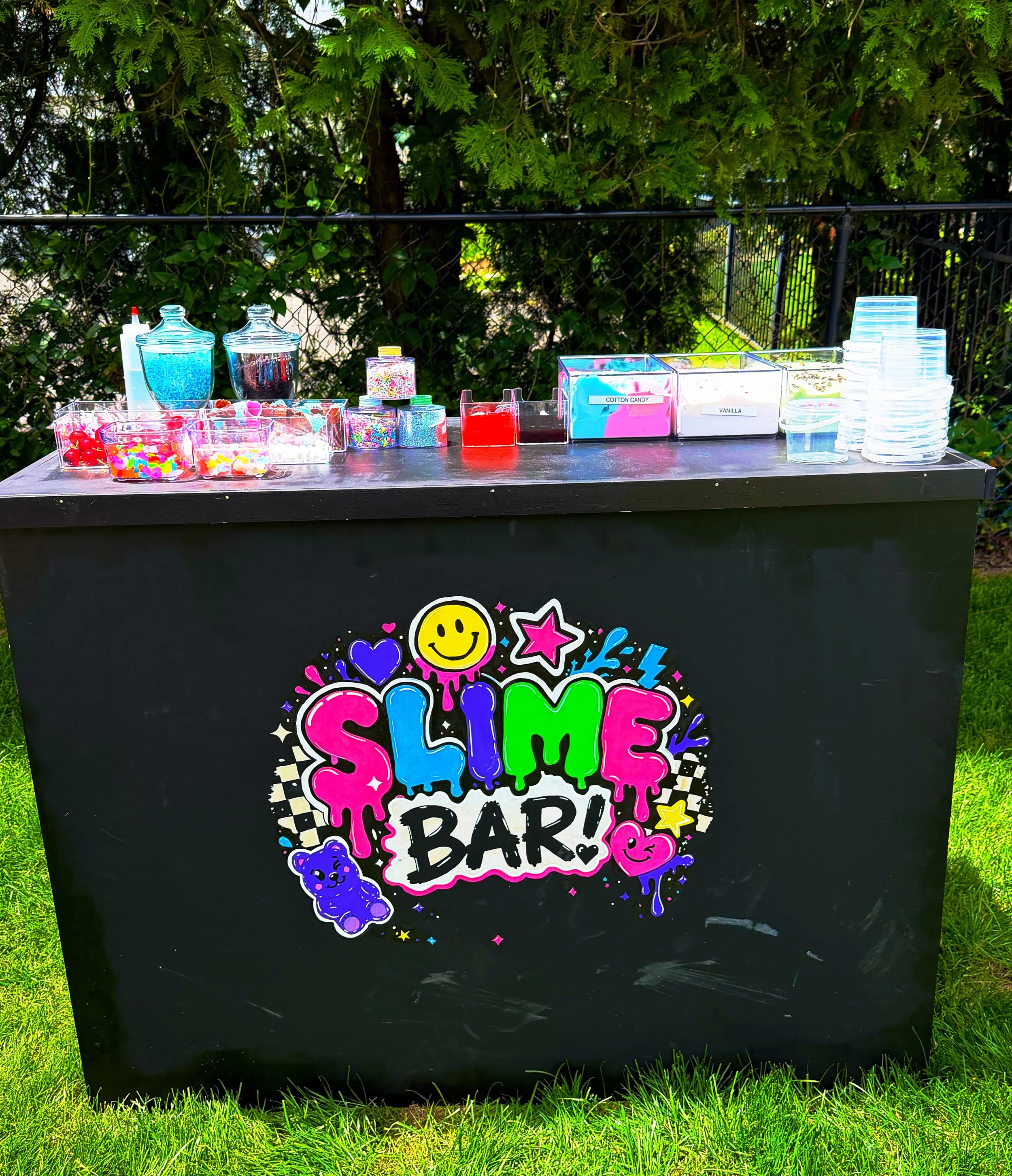 A black cart with colorful stickers that says 'Smile, It's My Birthday.' The cart is outdoors with a green grassy area and a black metal fence in the background, and the top of the cart is decorated with various containers of candy and snacks.