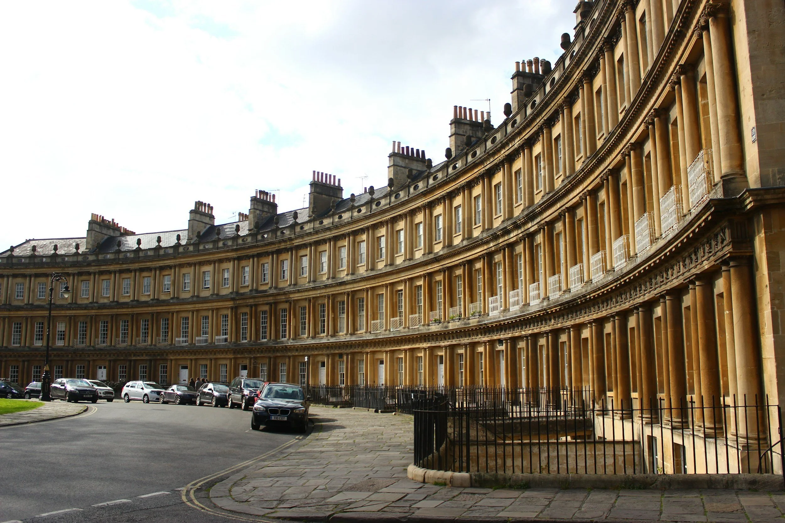 Curved row of historic yellow stone townhouses with multiple chimneys on rooftops, parked cars along the street, cloudy sky overhead.