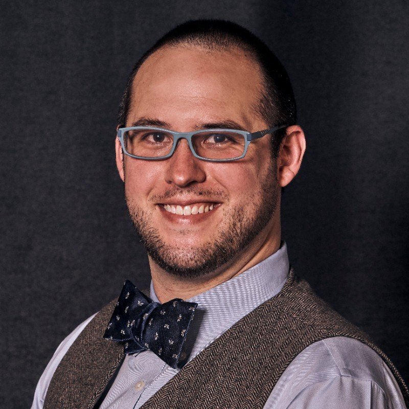 Portrait of a Eureka Northeast's founder and principal, Chris Maggiolo, with glasses, short dark hair, beard, wearing a bow tie, dress shirt, and vest against a dark background.