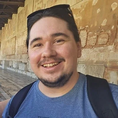 A young man with dark hair, a beard, and a moustache smiling outdoors, wearing sunglasses on his head, a blue T-shirt, and a backpack, with a stone wall in the background.