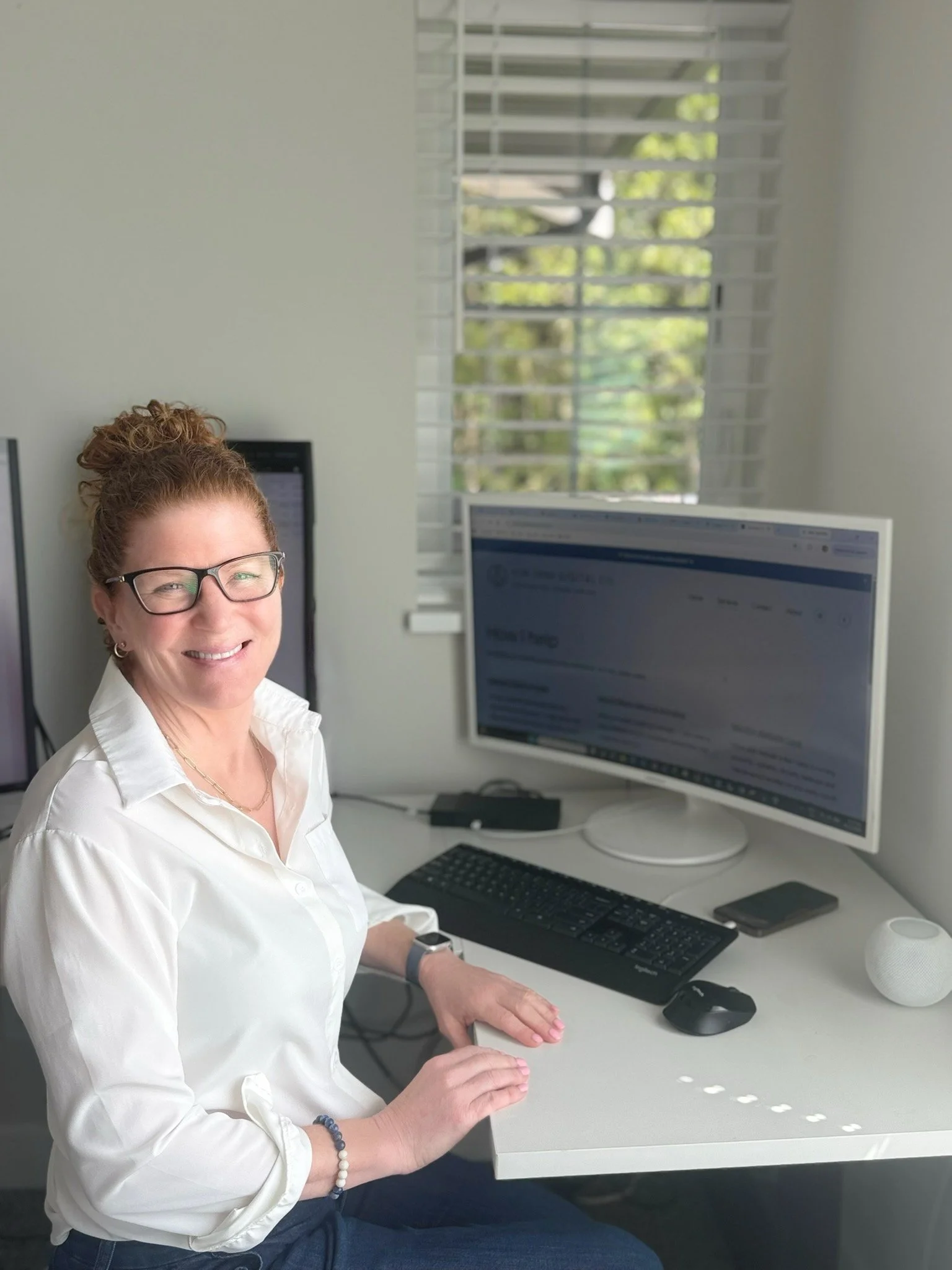 A woman in Gibsons BC, with glasses smiling at her desk with a computer monitor, keyboard, mouse, and smartphone in a bright room with large window blinds and greenery outside.