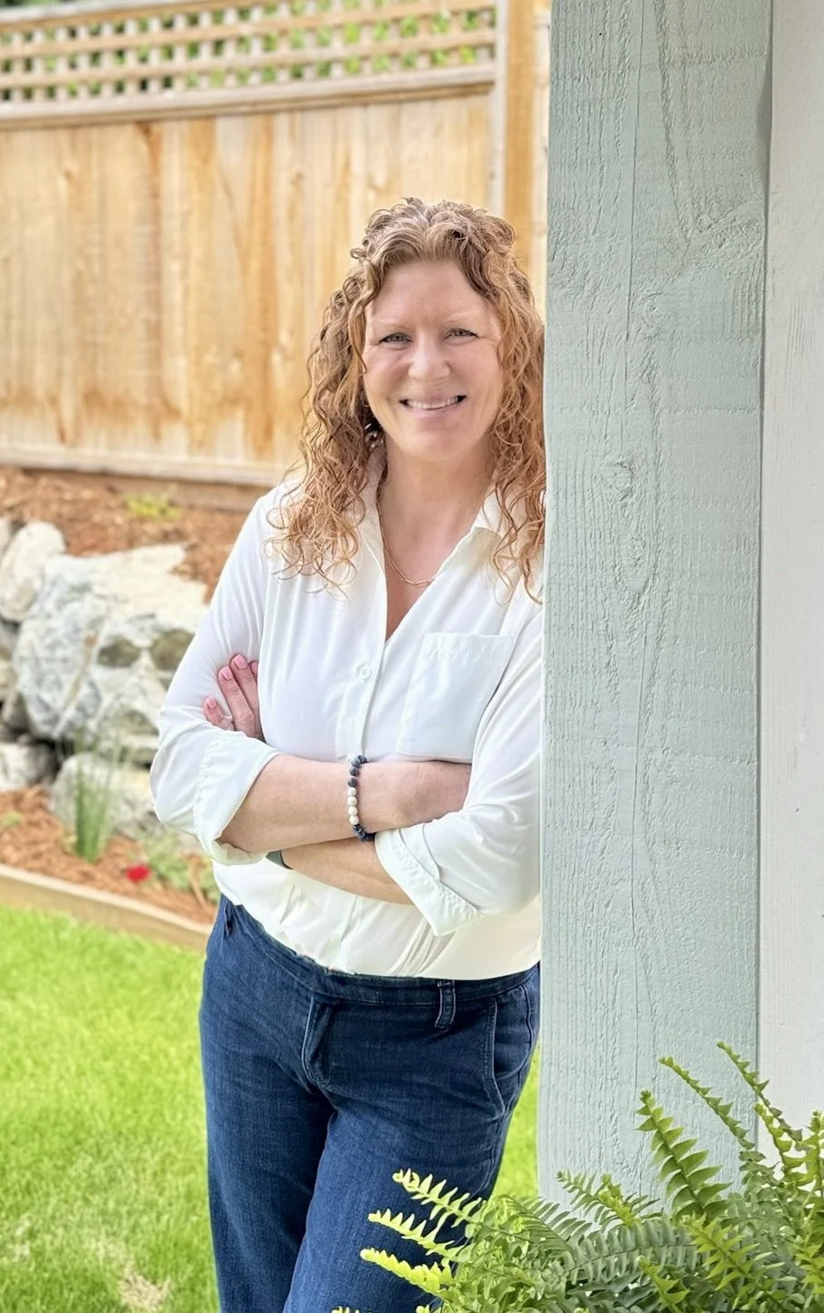 A woman in Gibsons BC with curly red hair smiling, standing outdoors next tall wooden post, arms crossed, with a wooden fence and garden in the background.