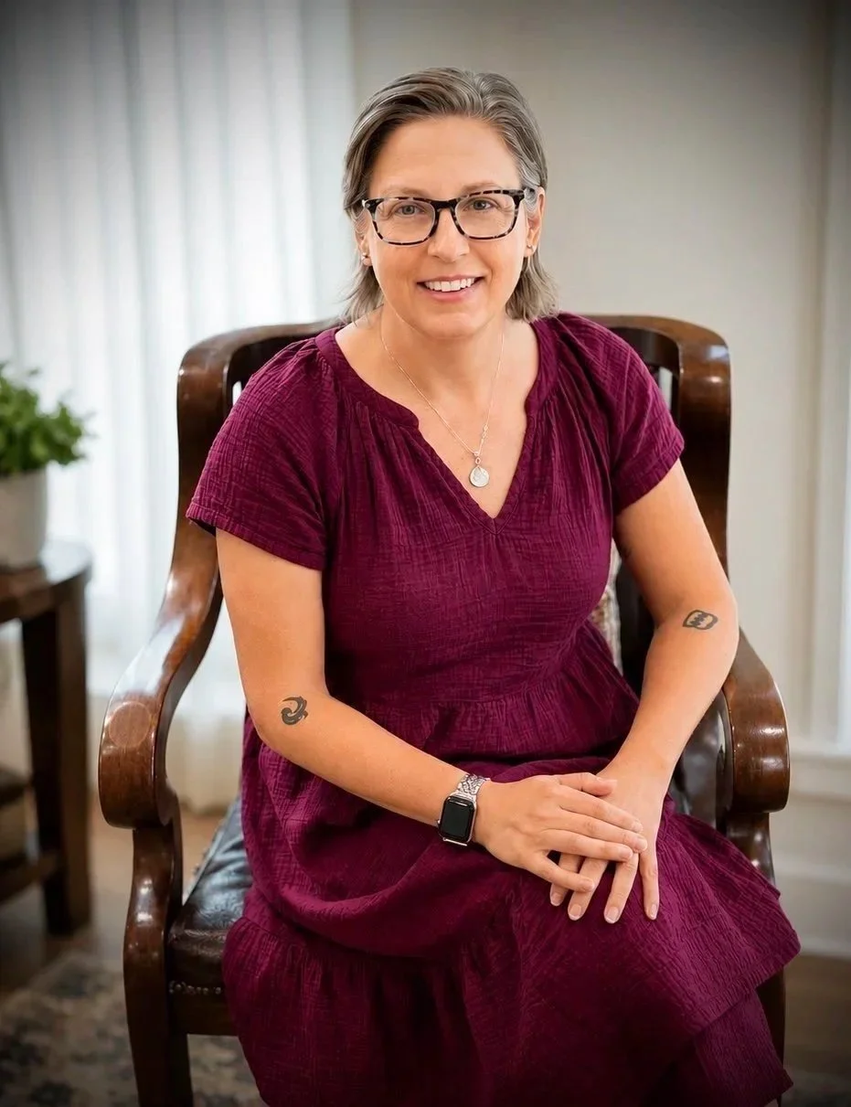 A woman with short gray hair and glasses, wearing a maroon dress, sitting on a wooden chair, smiling at the camera.