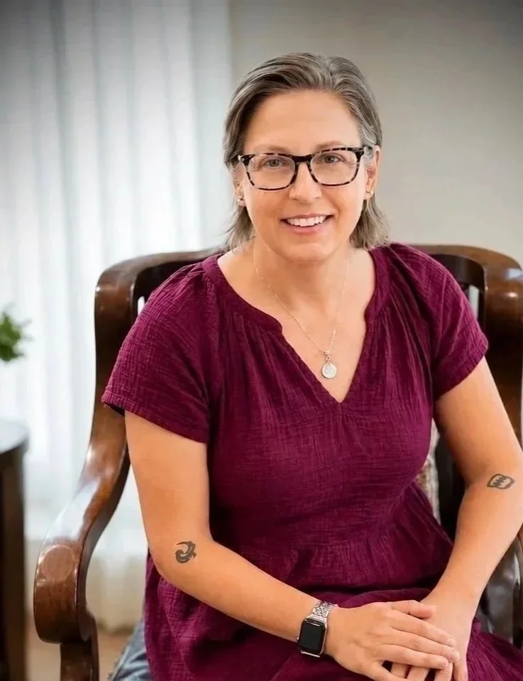A woman with shoulder-length gray hair, wearing glasses and a maroon short-sleeve dress, sitting on a wooden chair in a well-lit room with a smile.