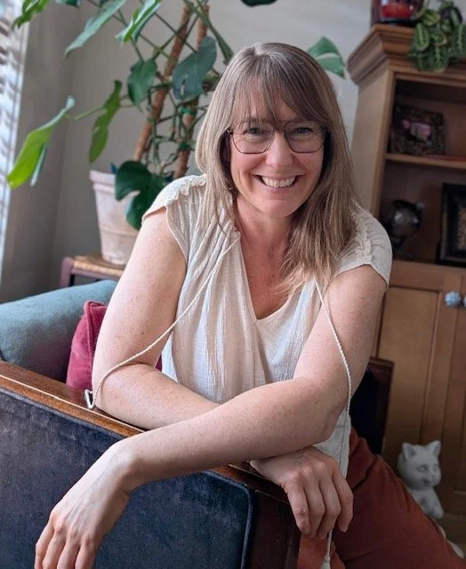 Woman with glasses and shoulder-length hair smiling, sitting at a wooden table in a cozy living room with plants and wooden furniture.