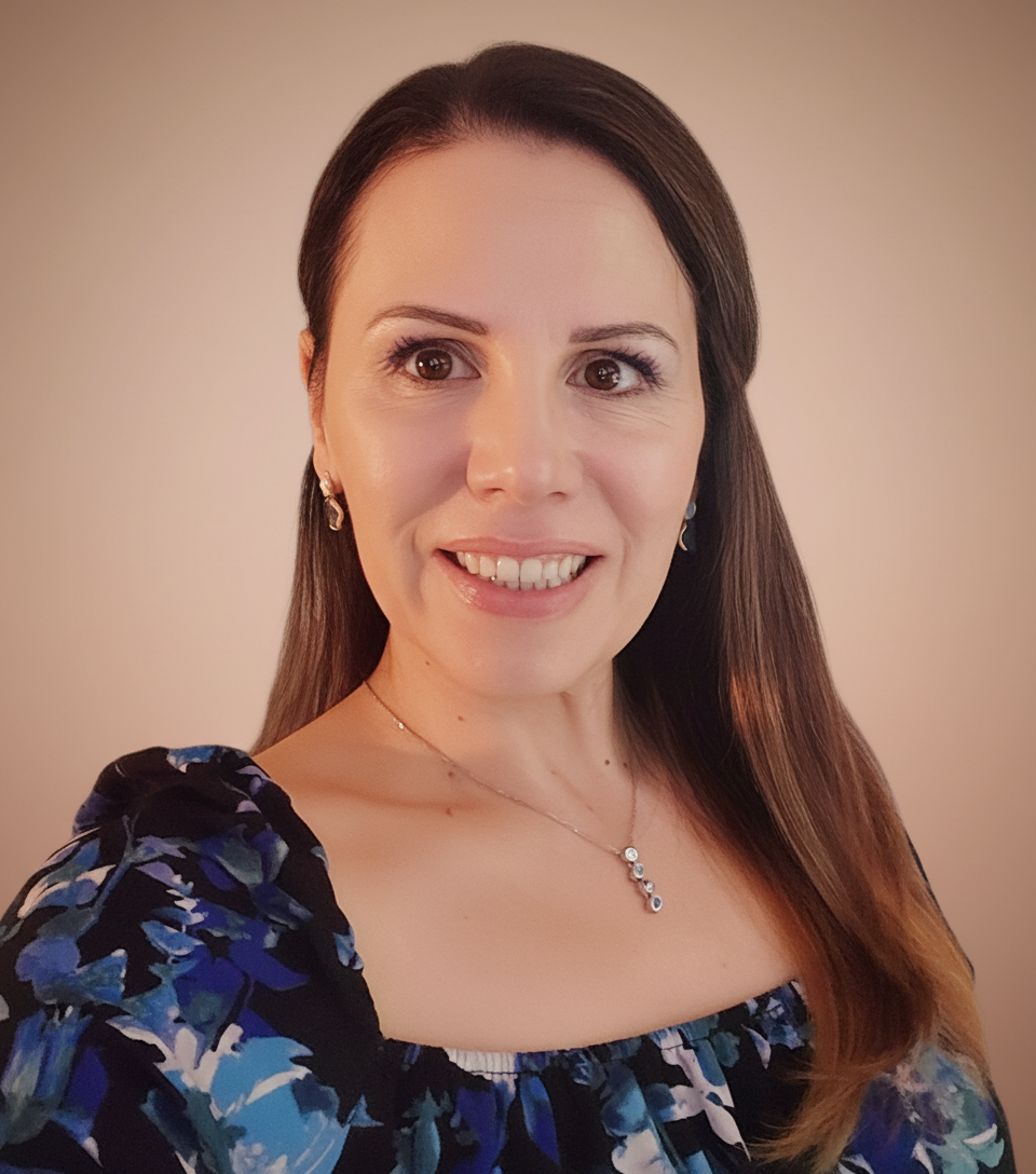 A woman with long brown hair and light skin smiling, wearing a floral dress with blue, black, and white colors, silver jewelry including earrings and a necklace, against a plain, light-colored background.