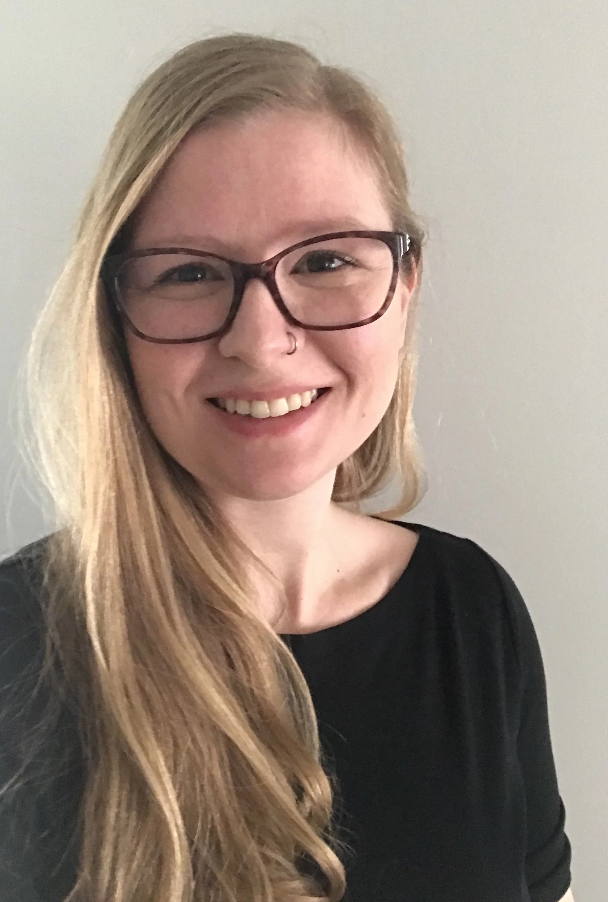 Close-up of a smiling woman with long, blonde hair, wearing black glasses and a nose ring, dressed in a black top, against a plain wall.