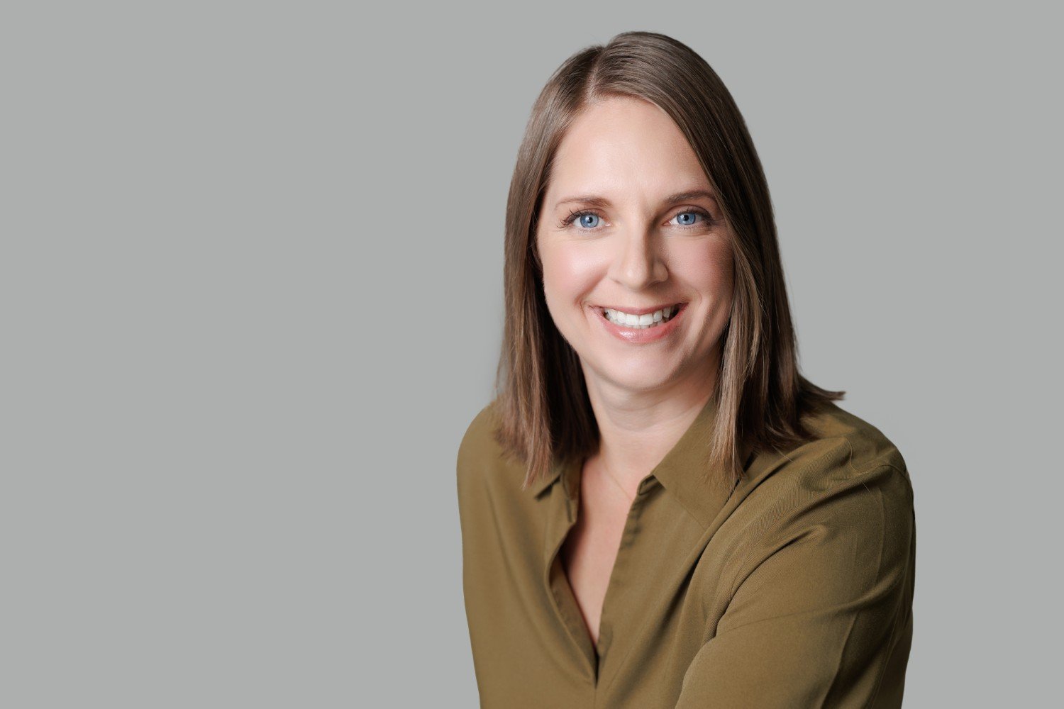A woman with shoulder-length brown hair, wearing a green blouse, smiling against a gray background.