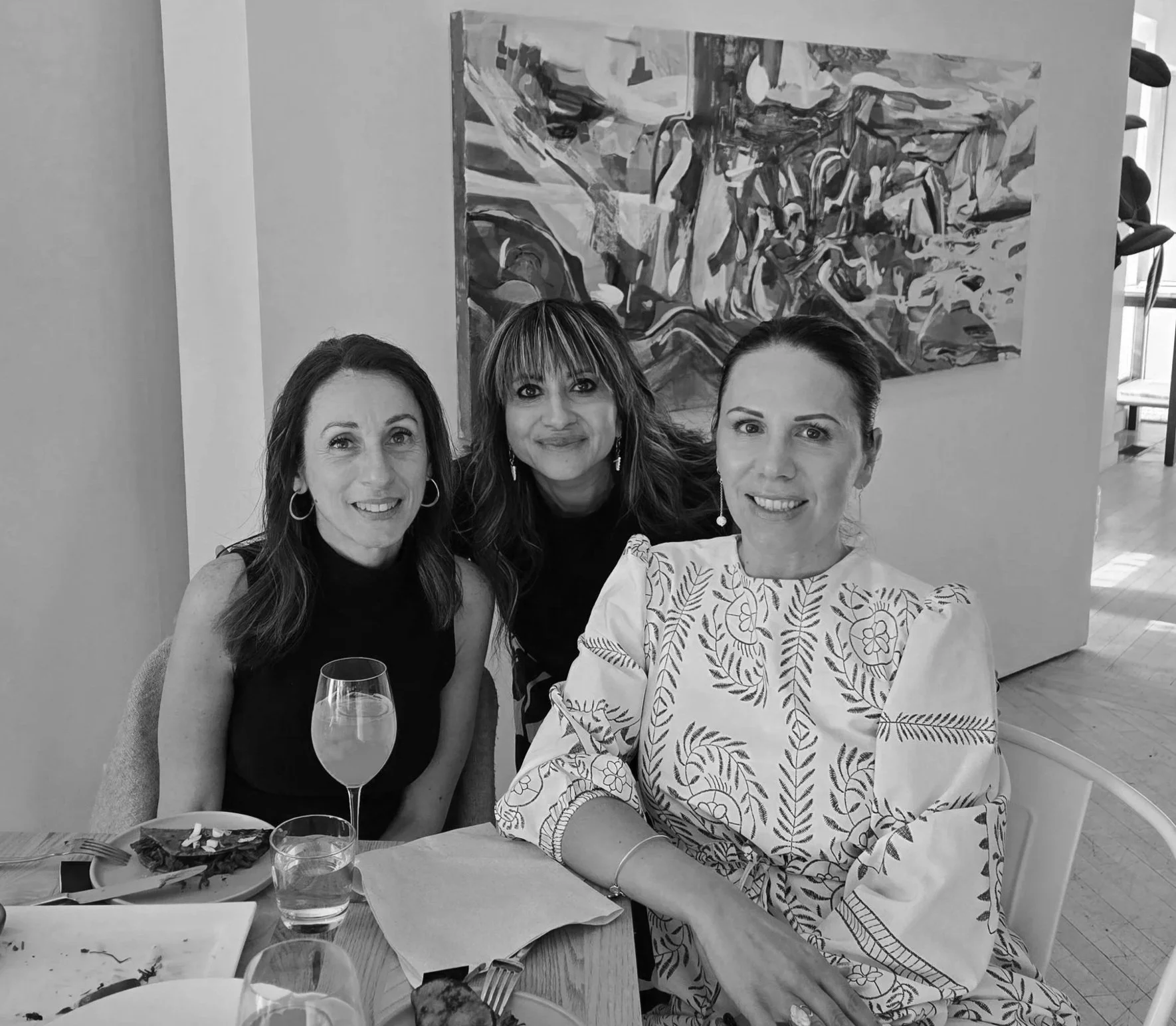 Three women sitting together at a dining table, smiling for a photo with a colorful abstract painting on the wall behind them. There are drinks and a plate of food on the table.