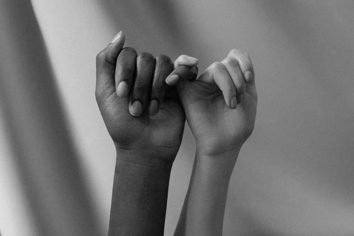 Two hands, one dark-skinned and one light-skinned, holding each other with pinky fingers intertwined.