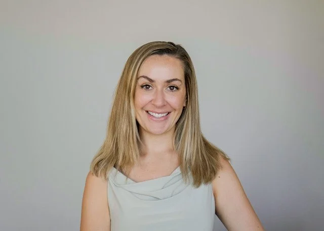 A smiling woman with blonde hair wearing a light sleeveless top, standing against a plain light-colored background.