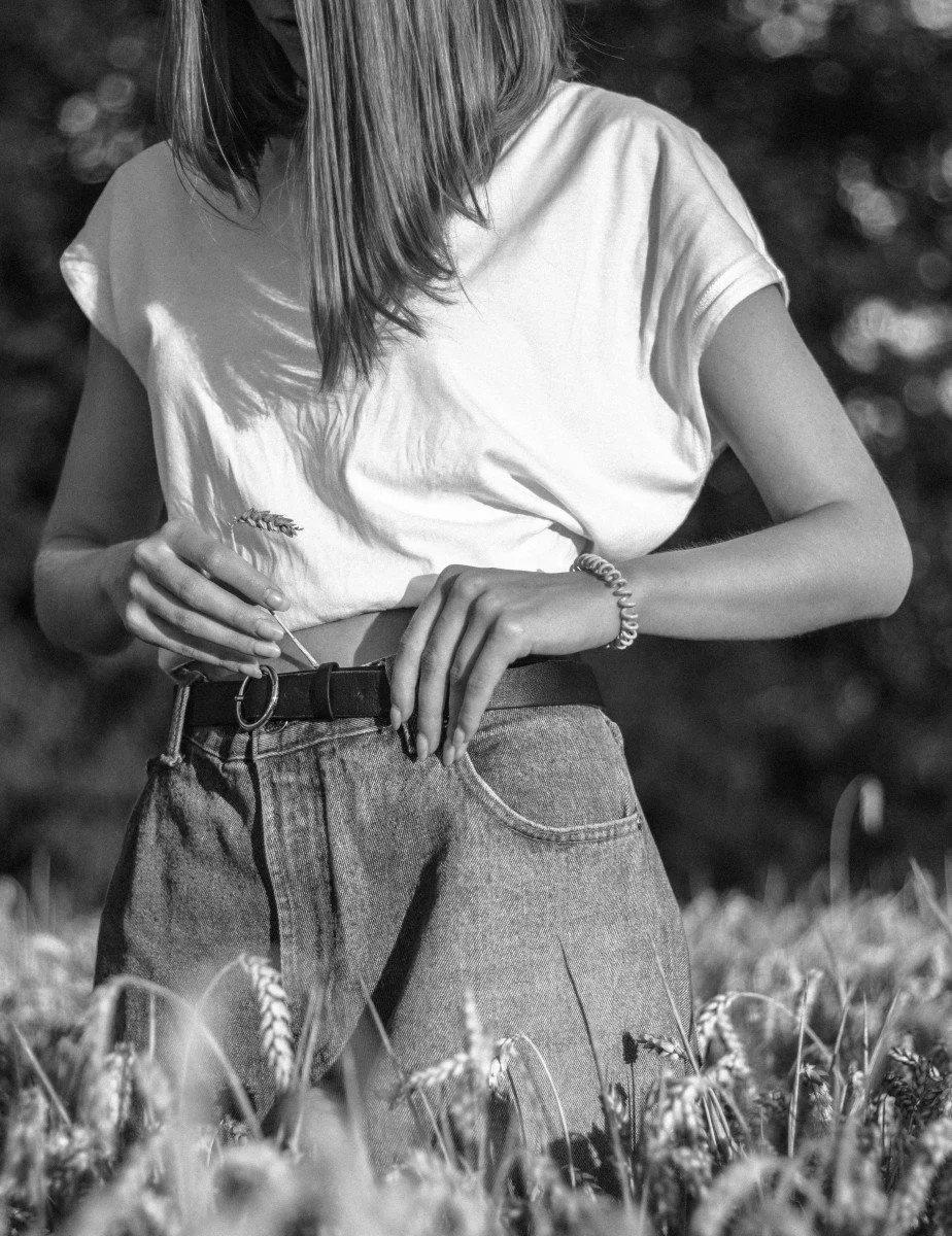 A person wearing a white t-shirt and jeans standing in a wheat field, holding a wheat stalk with one hand.