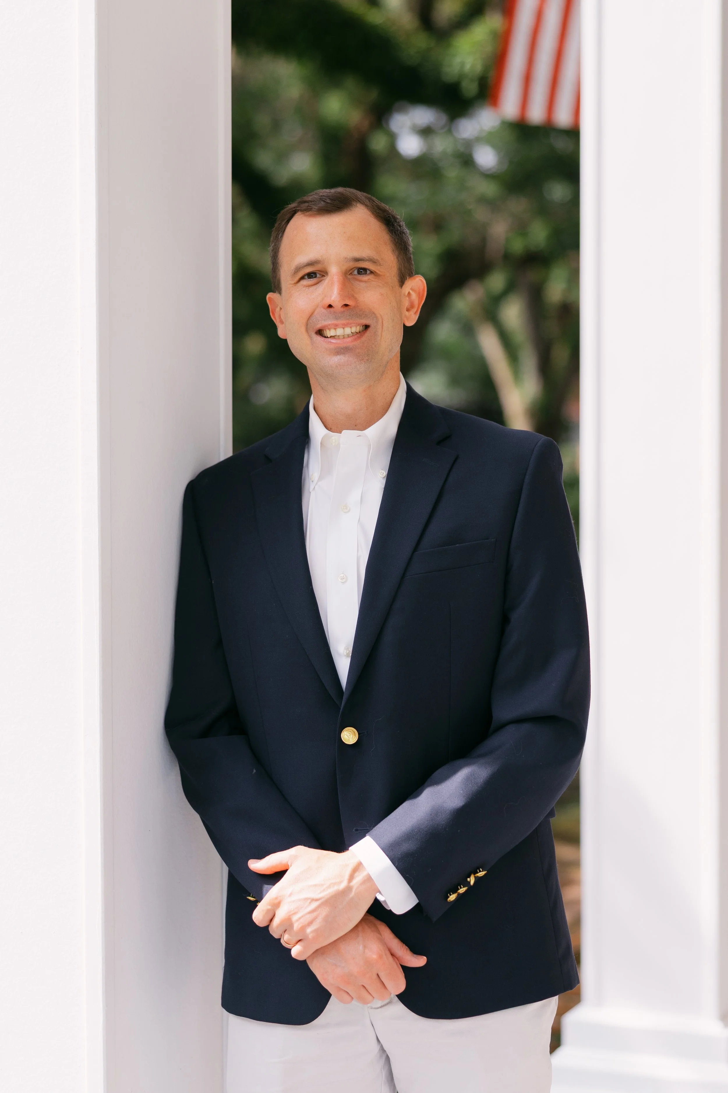 A man in a navy blazer and white shirt is standing outdoors by a white wall, smiling, with trees and an American flag in the background.