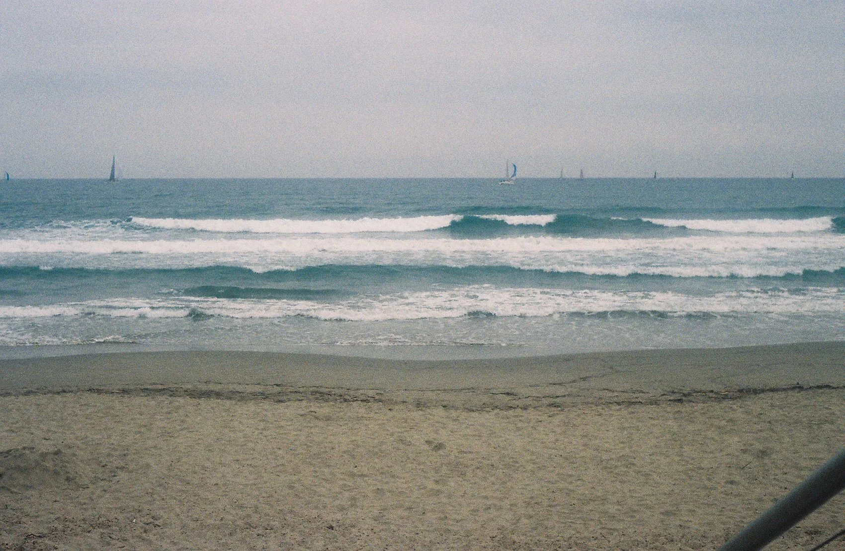 A sandy beach with gentle ocean waves and sailboats on the horizon under a gray sky.