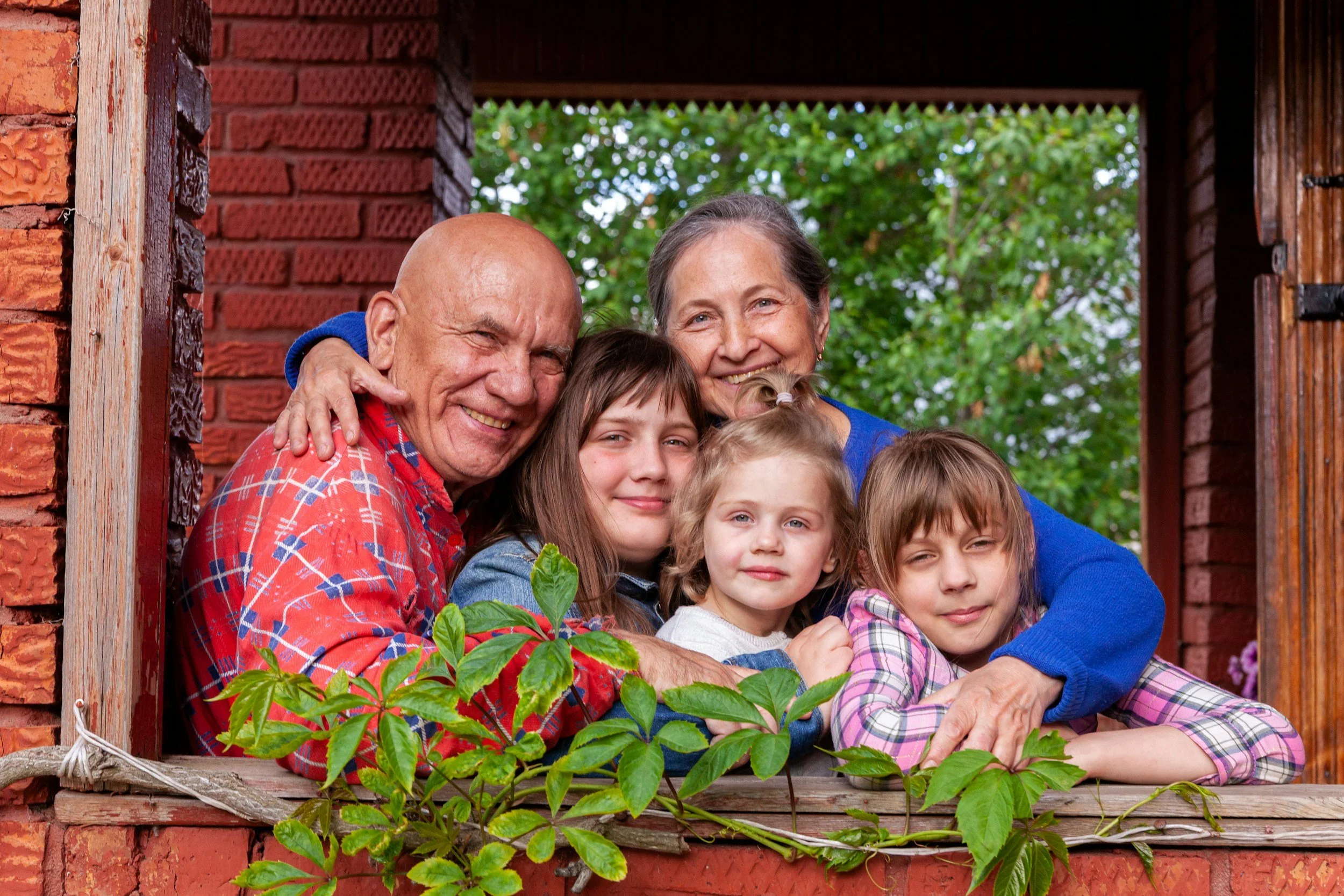 A happy multigenerational family of five, including an elderly man and woman, two young girls, and a young boy, smiling and hugging each other on a wooden porch with brick walls and green foliage in the background.