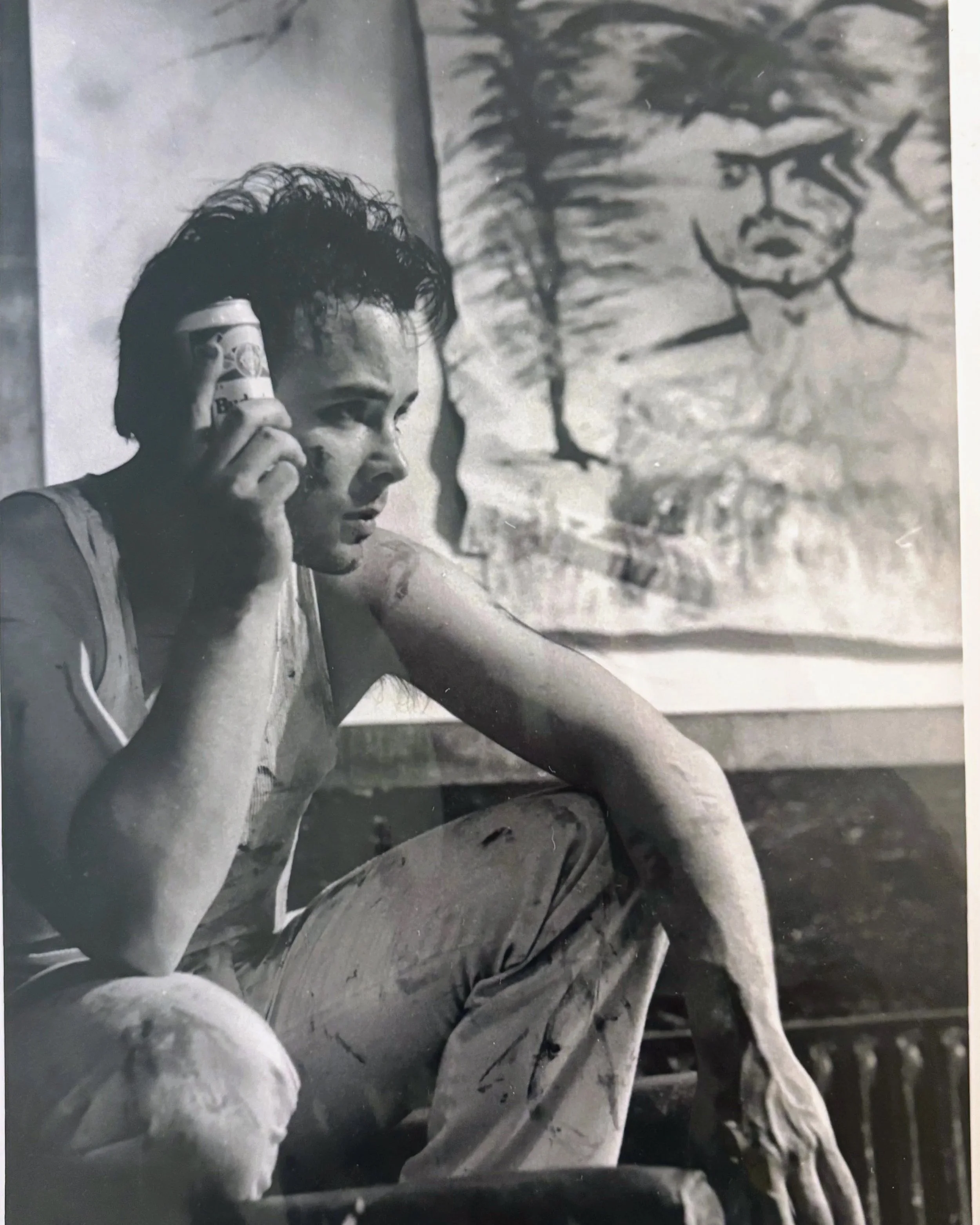 A young man with messy, curly hair is talking on a cell phone and looking off into the distance. In the background, there is a large painting of a man with glasses and a beard, giving a serious expression.