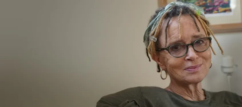 A middle-aged woman with glasses and multicolored dreadlocks, wearing a dark shirt, sitting in a room with a neutral-colored wall and artwork in the background.