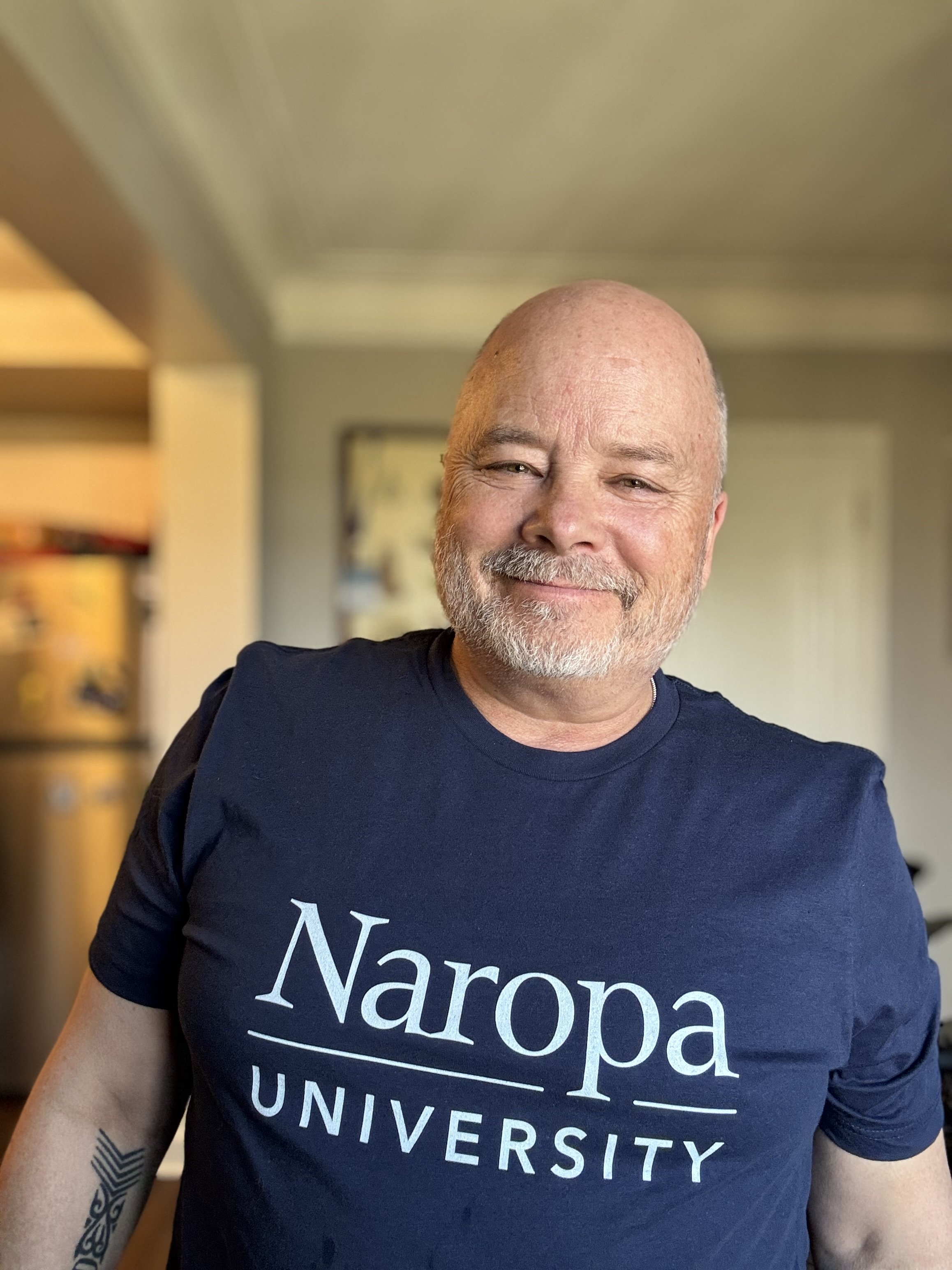A smiling man with a beard and tattoos on his arm wearing a navy blue t-shirt with white text that says 'Naroa University'.