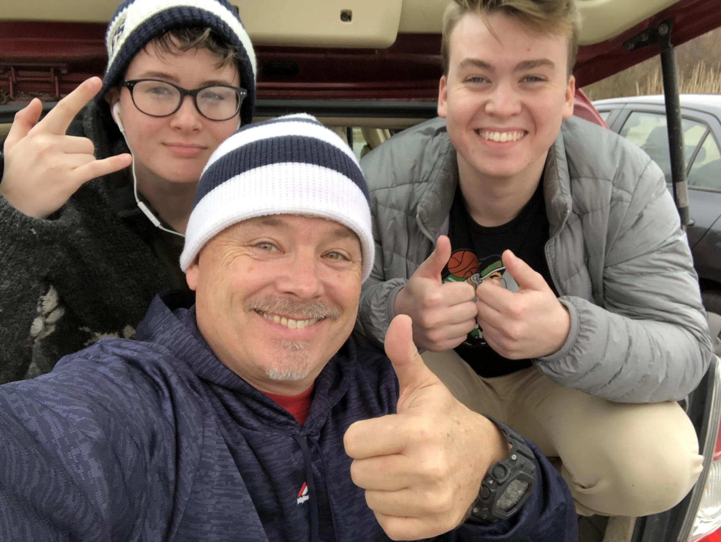Four people smiling and giving thumbs up indoors, with a vehicle in the background.