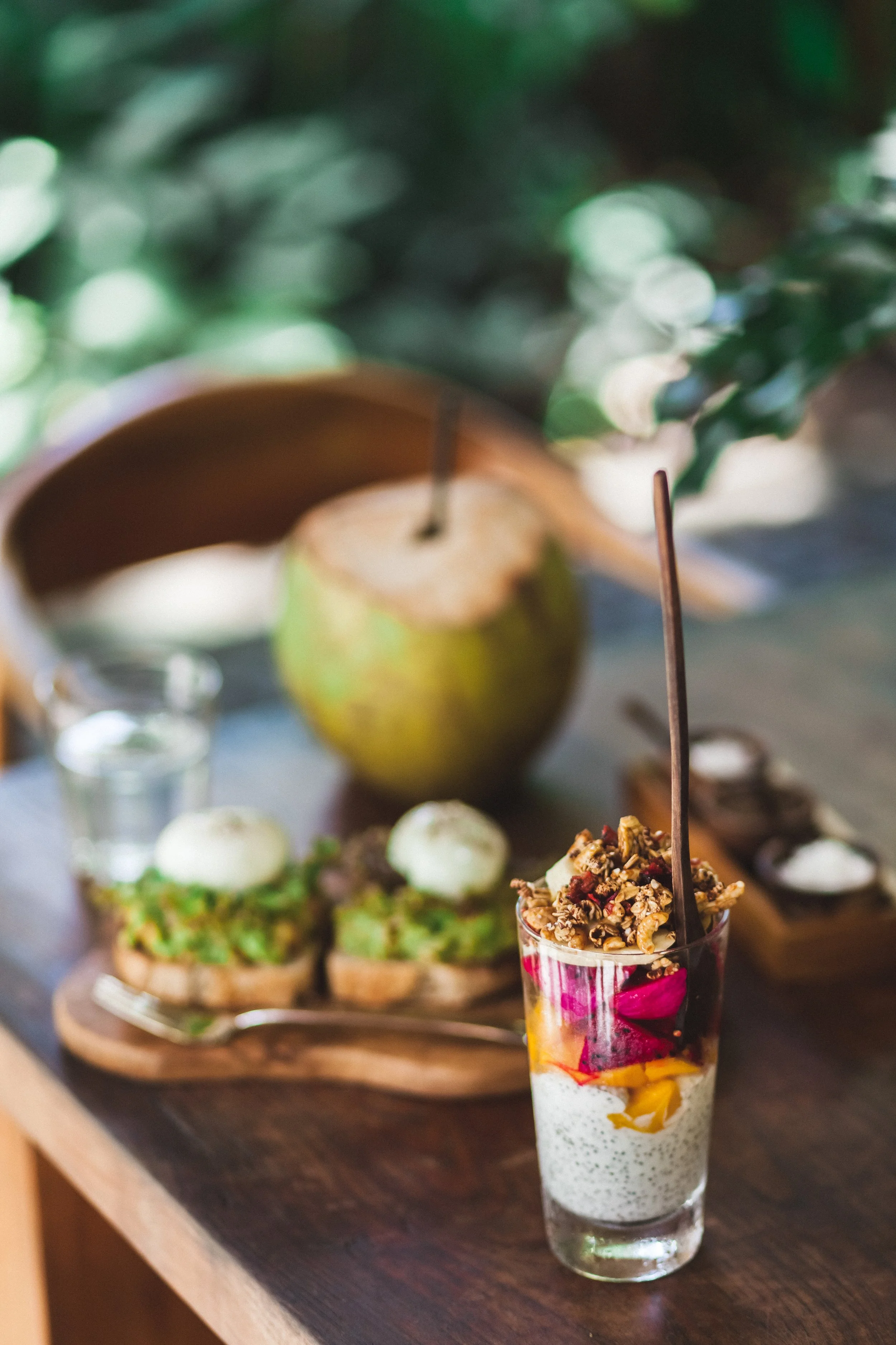 A layered dessert in a tall glass with various fruit and whipped cream on top, placed on a dark wooden table.