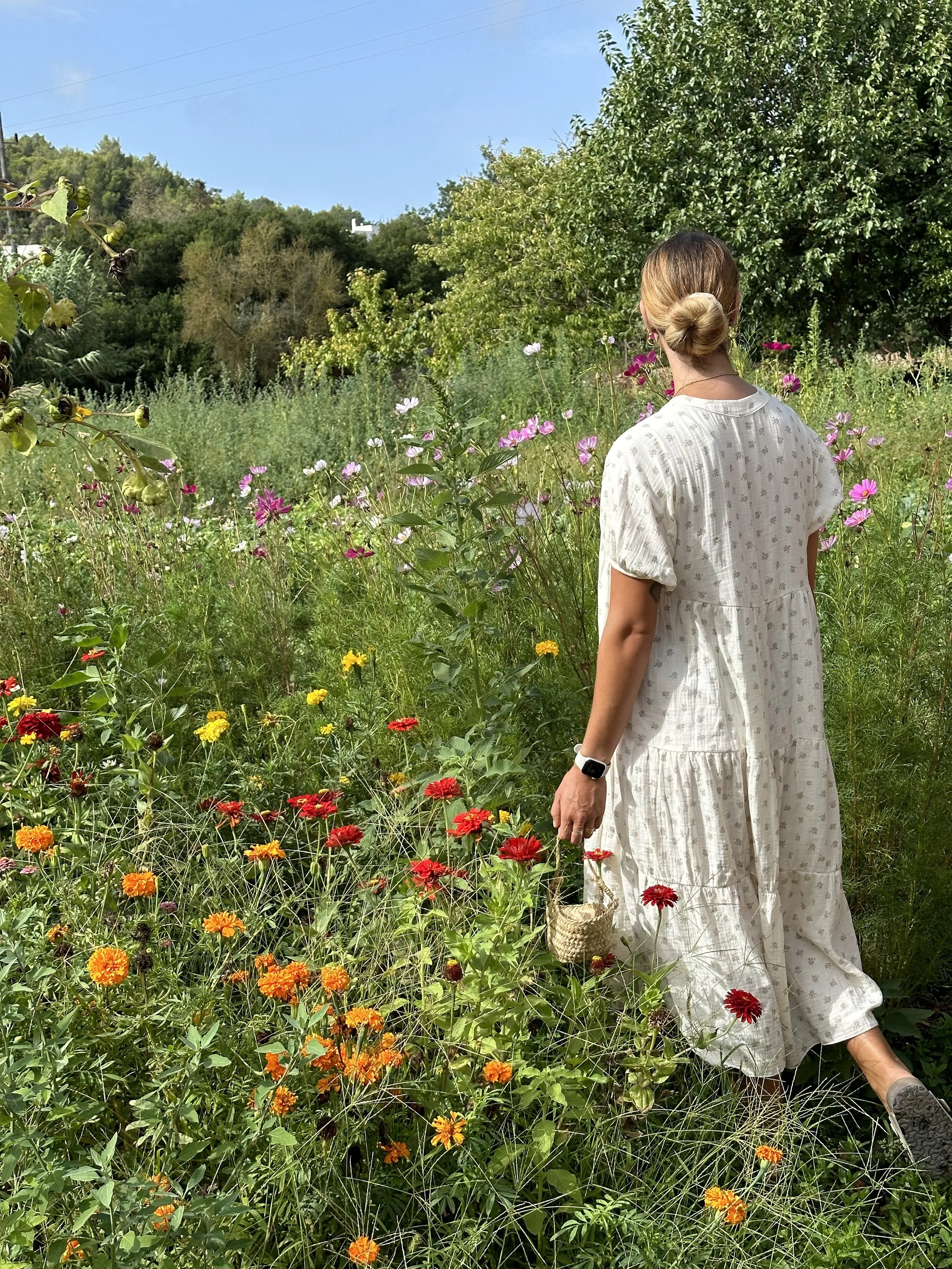 A woman with light brown hair tied in a bun, wearing a white floral dress, walking through a garden filled with colorful flowers including red, orange, pink, and white blooms, holding a small woven basket, with green trees and a blue sky in the background.