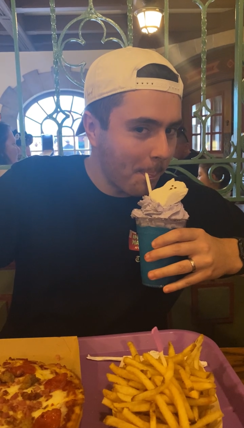 A young man wearing a white baseball cap backwards, drinking a purple ice cream float with a ghost-shaped marshmallow, seated at a table with pizza and French fries in a restaurant.