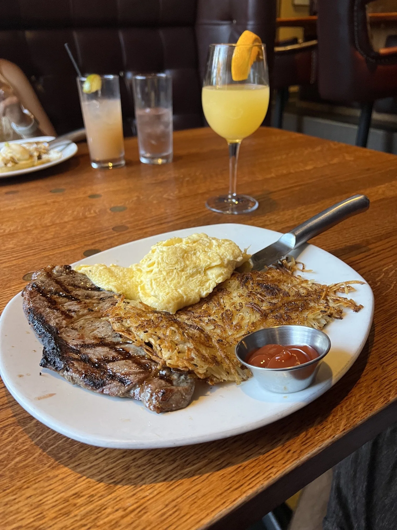 A plate with a large grilled beef steak, scrambled eggs, hash browns, and a small cup of ketchup. In the background, there are three glasses of drinks, one with a lemon wedge, on a wooden table in a restaurant setting.