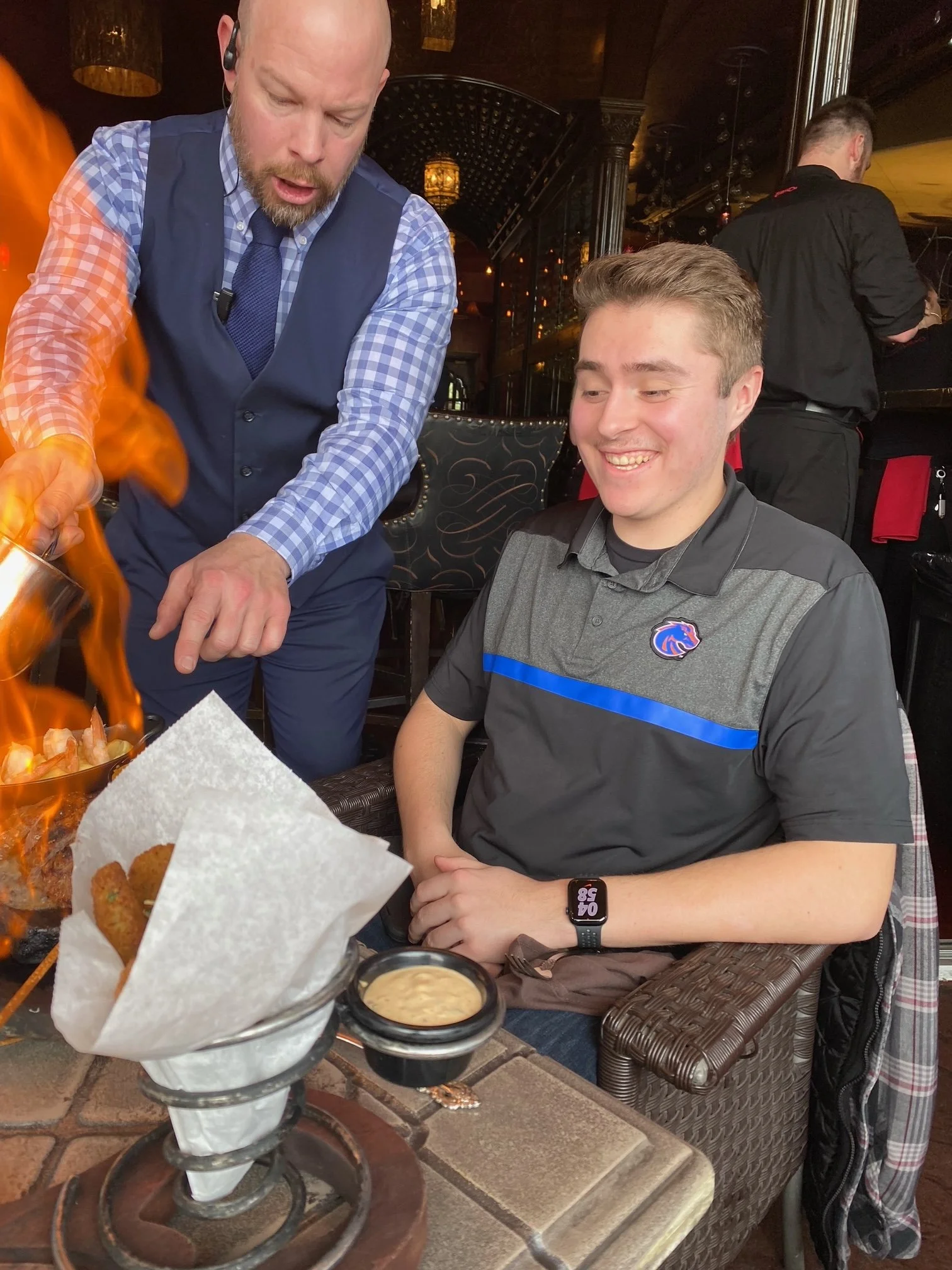A man with short light brown hair and a gray shirt with blue stripes, sitting at a restaurant table, smiling as a waiter presenting food with flames. The waiter is wearing a checkered shirt, a blue vest, and a tie. In the background, another person with short hair is standing, wearing a black shirt. The table has a basket with fried foods, dipping sauces, and a candle.