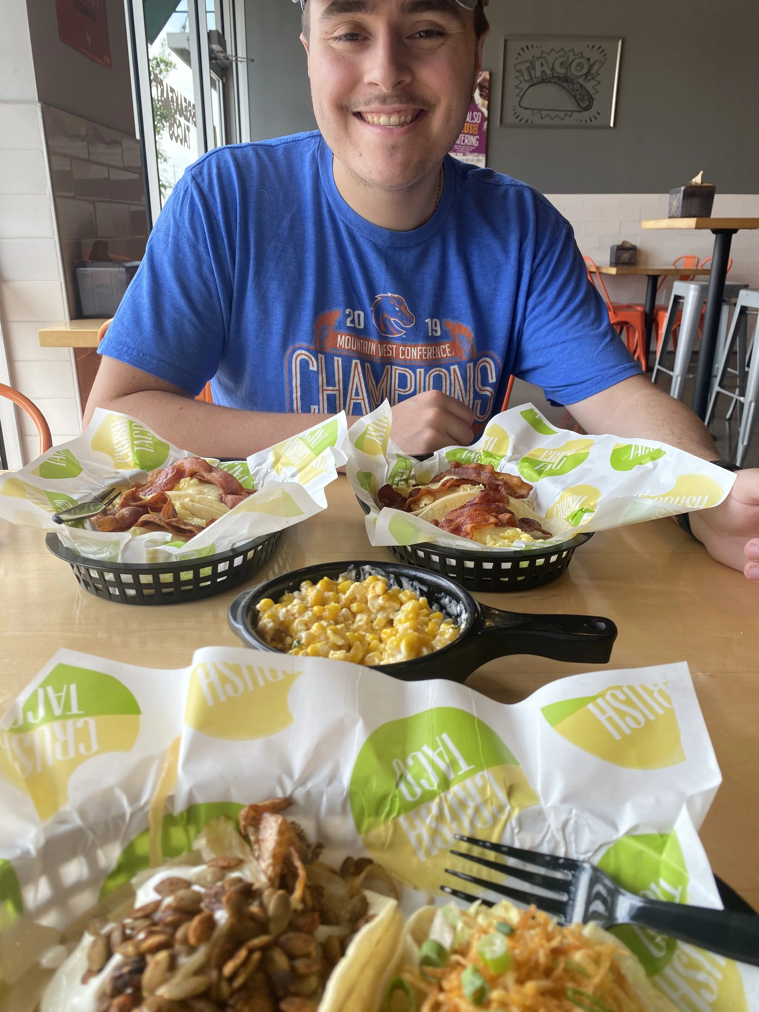 A young man smiling at the camera, sitting at a table with two baskets of breakfast food, including bacon, eggs, and hash browns, in a casual restaurant setting.