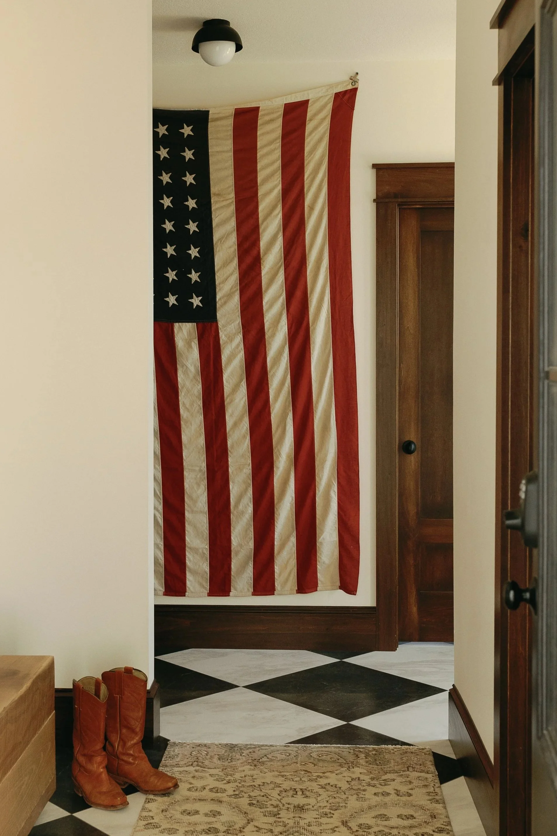 An American flag hanging in a hallway near a wooden door with a black knob. There are cowboy boots and a pair of shoes on the floor near a beige rug with a floral pattern. The ceiling has a black and white ceiling light. The floor is checkered with b