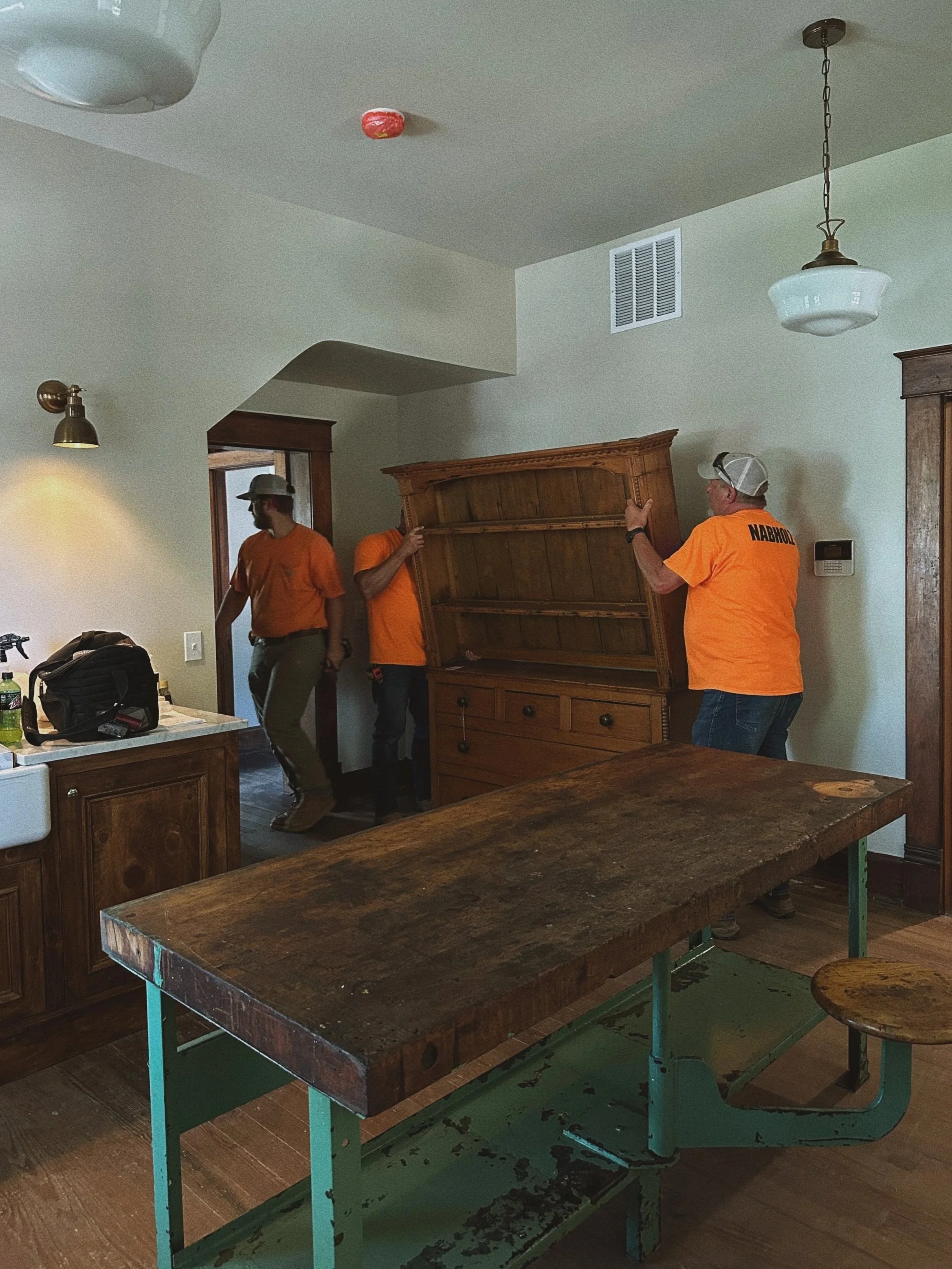 Three men in orange shirts moving a large wooden hutch inside a house. One man is helping at the front, another man on the side, and the third man is near the doorway.