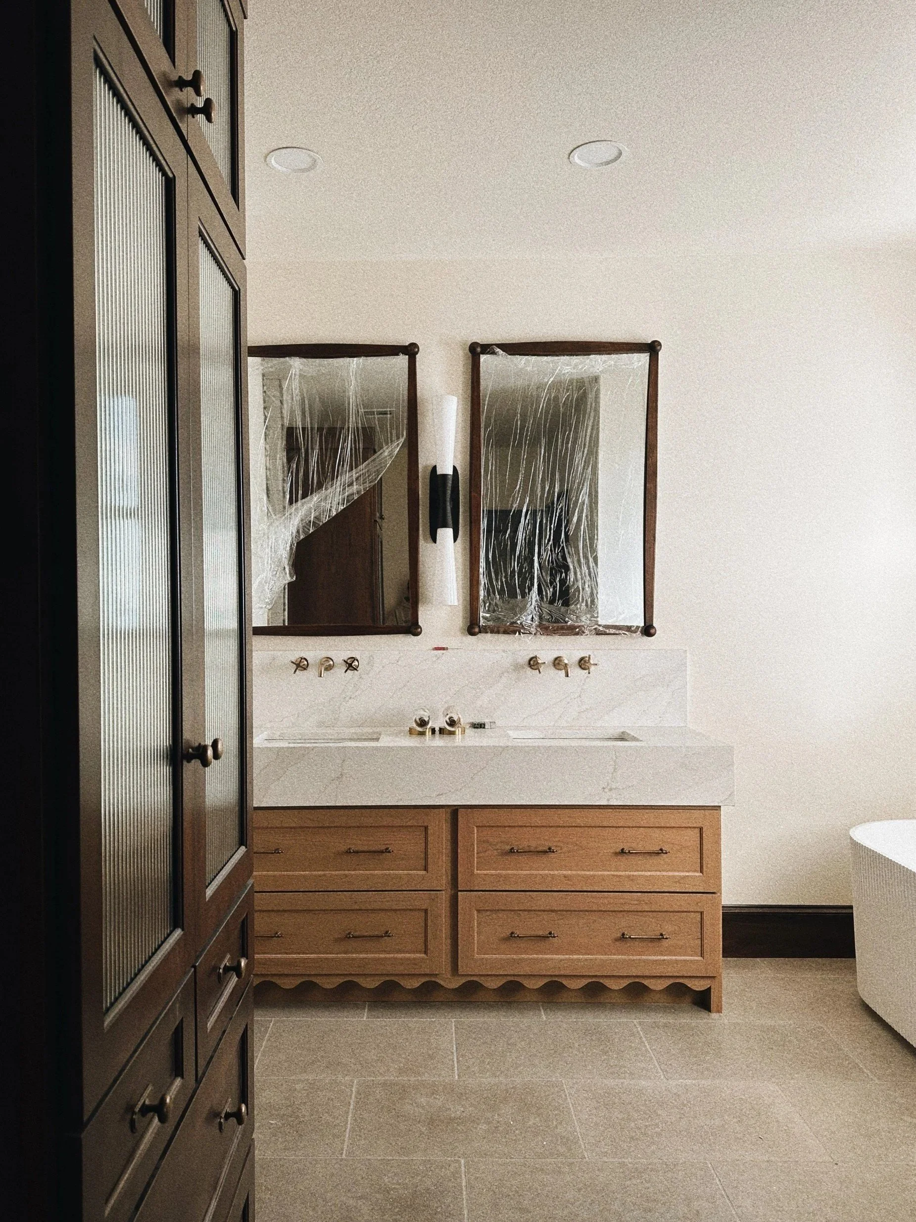 Bathroom vanity with two mirrors, a white countertop, brass faucets, a dark wooden cabinet, and a beige wall with two ceiling lights.
