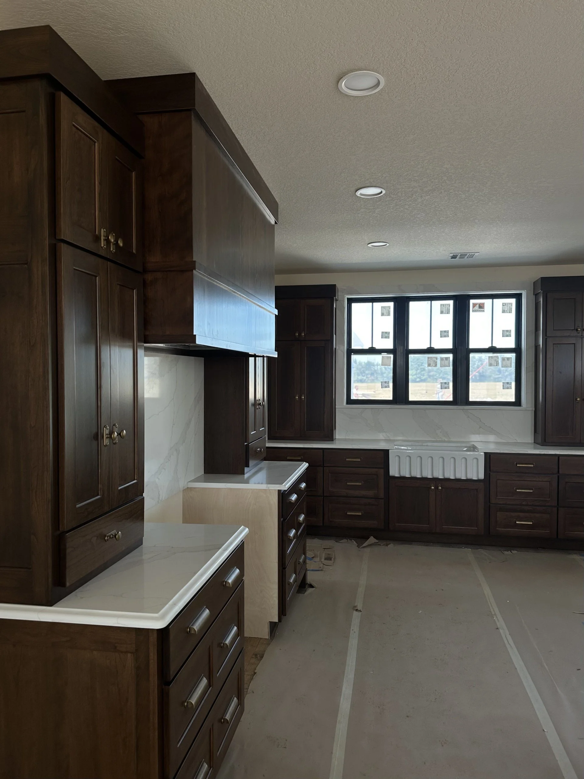 Kitchen under construction with dark wood cabinets, white countertops, and a large window, showing flooring protection and construction materials.