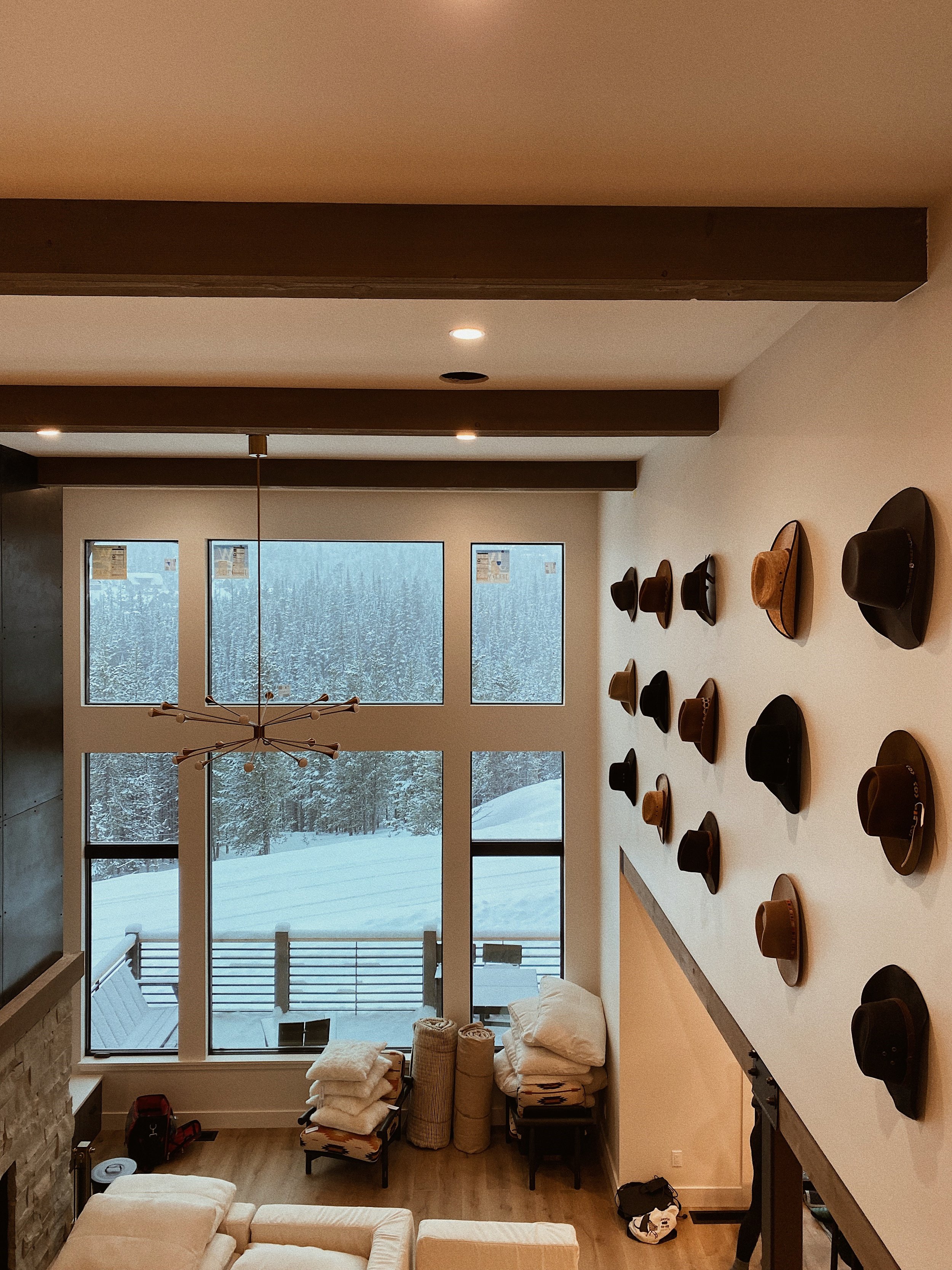 Modern living room with large windows overlooking snowy landscape, ceiling beams, contemporary chandelier, wall-mounted hats, and cozy furniture.