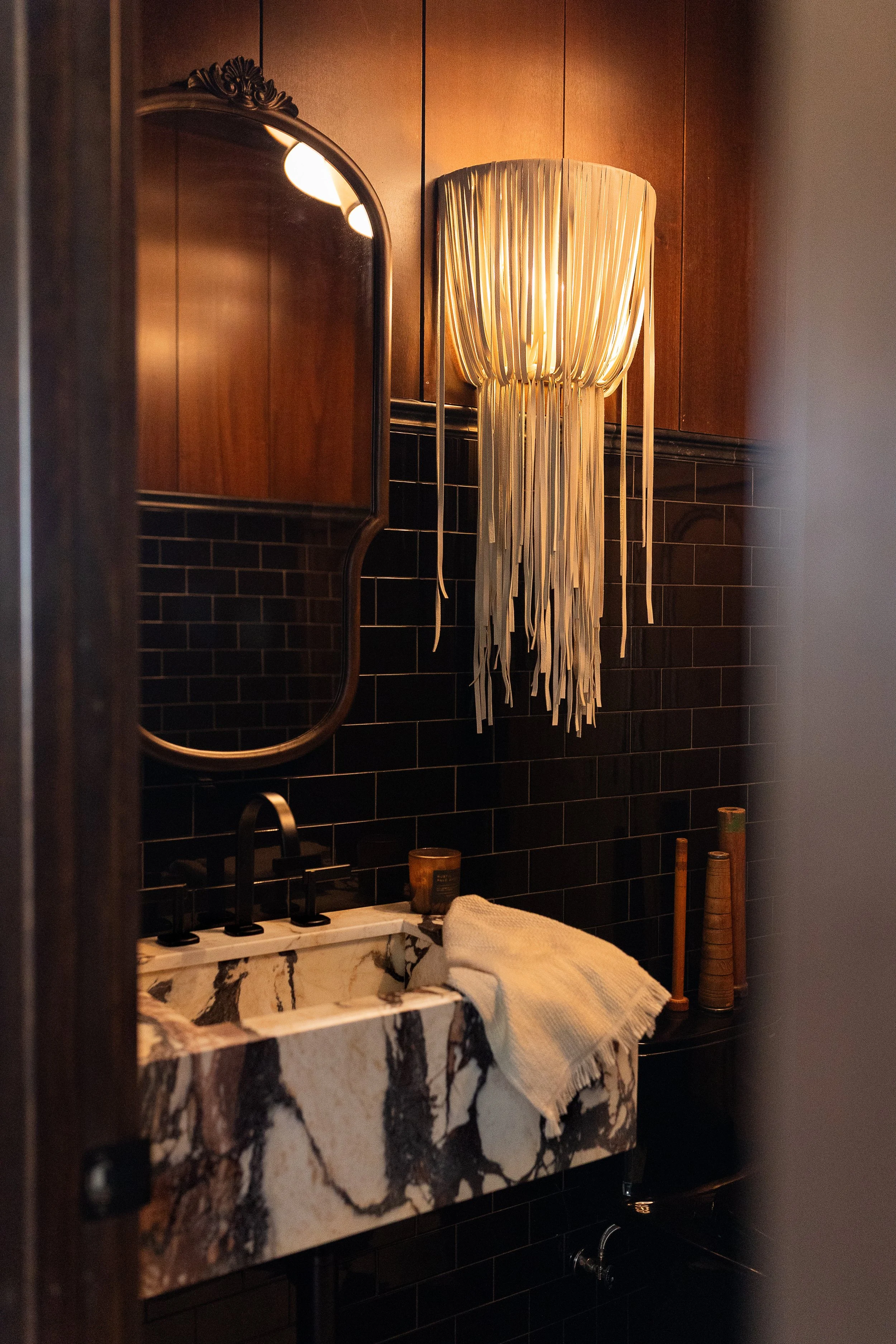 A bathroom with a marble sink, black faucet, and black tiled walls. A wall-mounted lamp with beige drapes and ribbons hangs above the sink, and a mirror reflects part of the room. There are some bamboo objects beside the sink, and a towel draped over