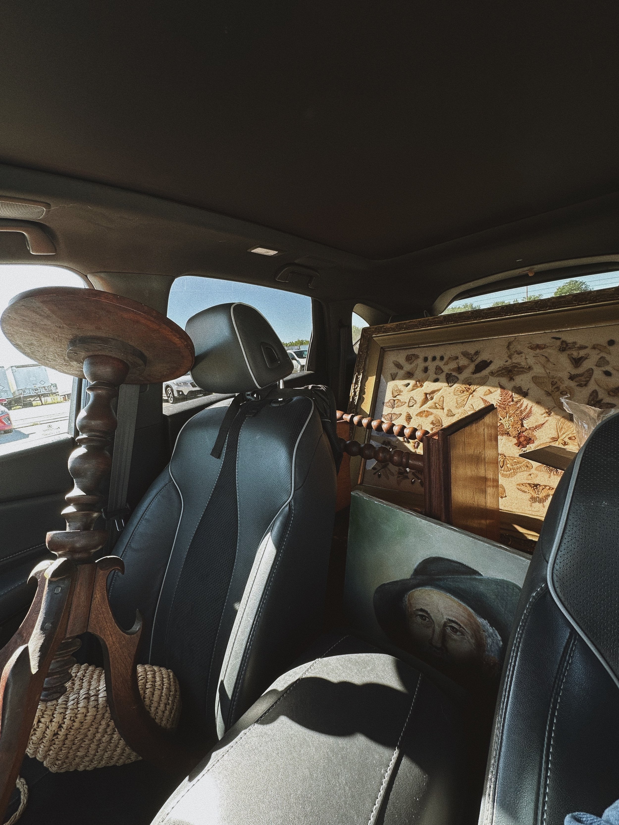 Inside a vehicle, filled with various objects, including a wooden spinning wheel, framed artwork, and furniture with a light beige seat.