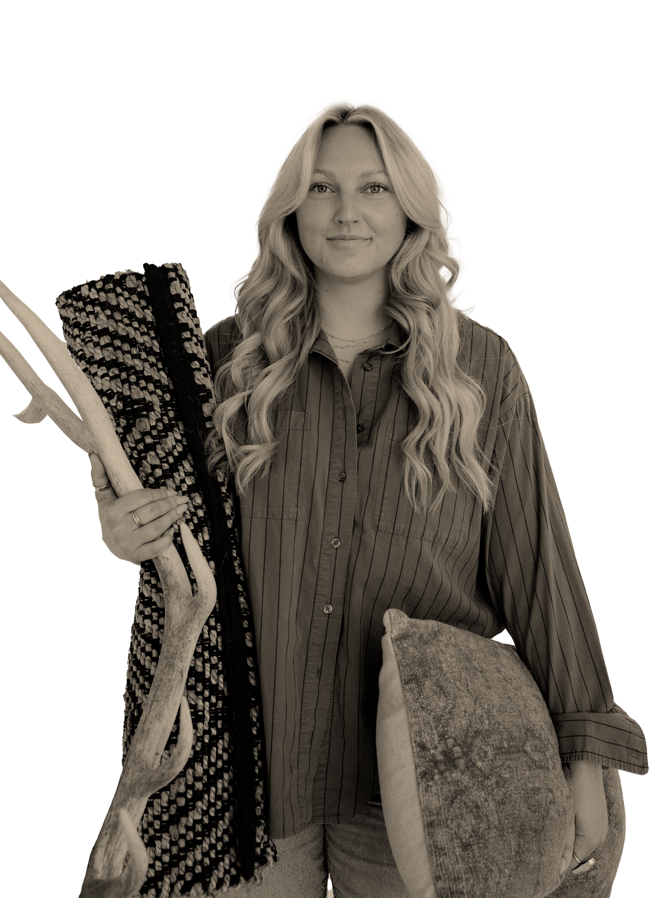 A woman with long wavy blonde hair holding a large patterned blanket and a piece of wood, dressed in a striped button-up shirt, standing with a large textured pillow in front of her.
