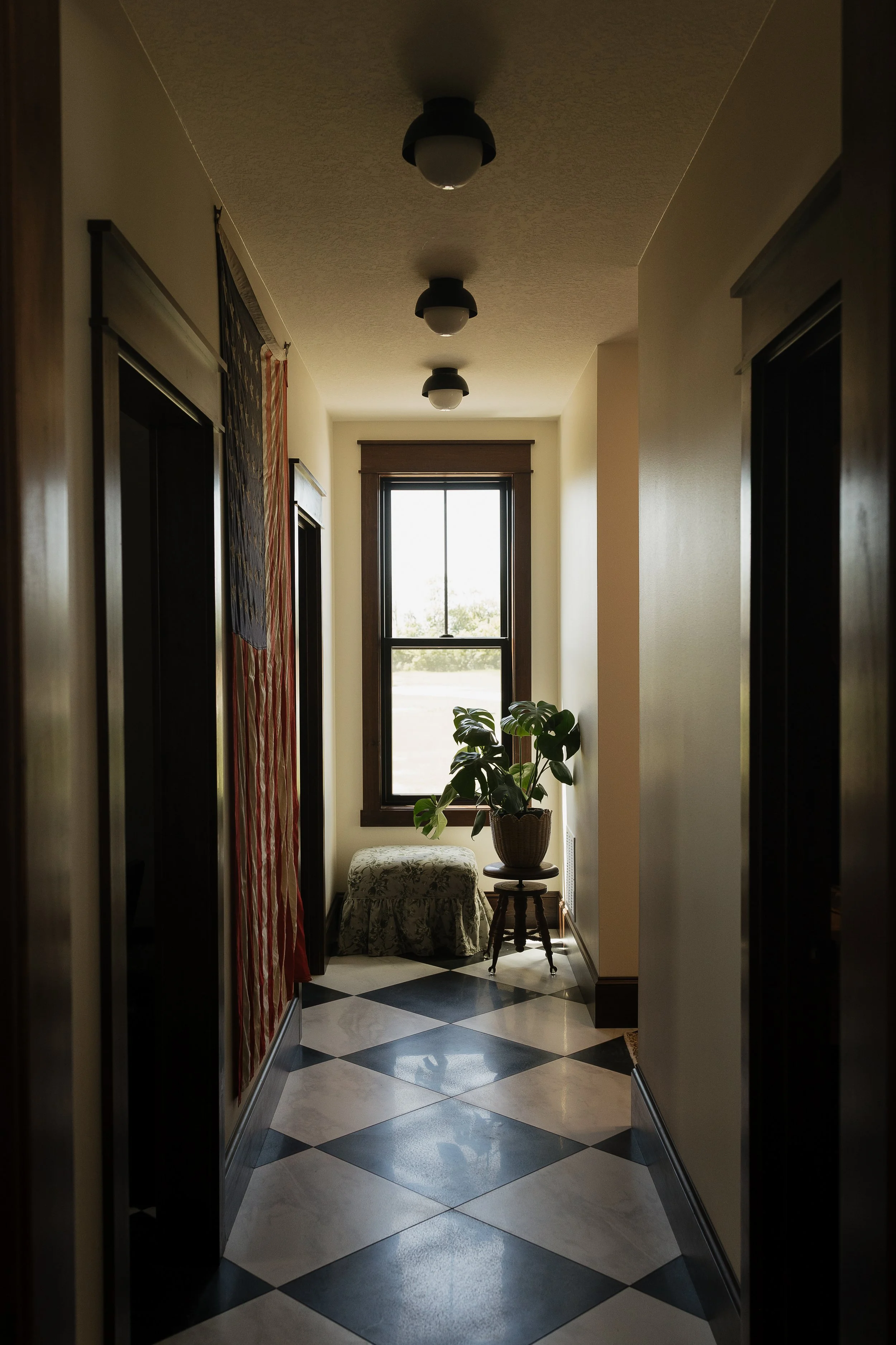 A hallway with a black and white checkered floor, cream-colored walls, and a window at the end. There is a potted plant on a stand in front of the window, and a small decorated ottoman. The ceiling has three black and white light fixtures.