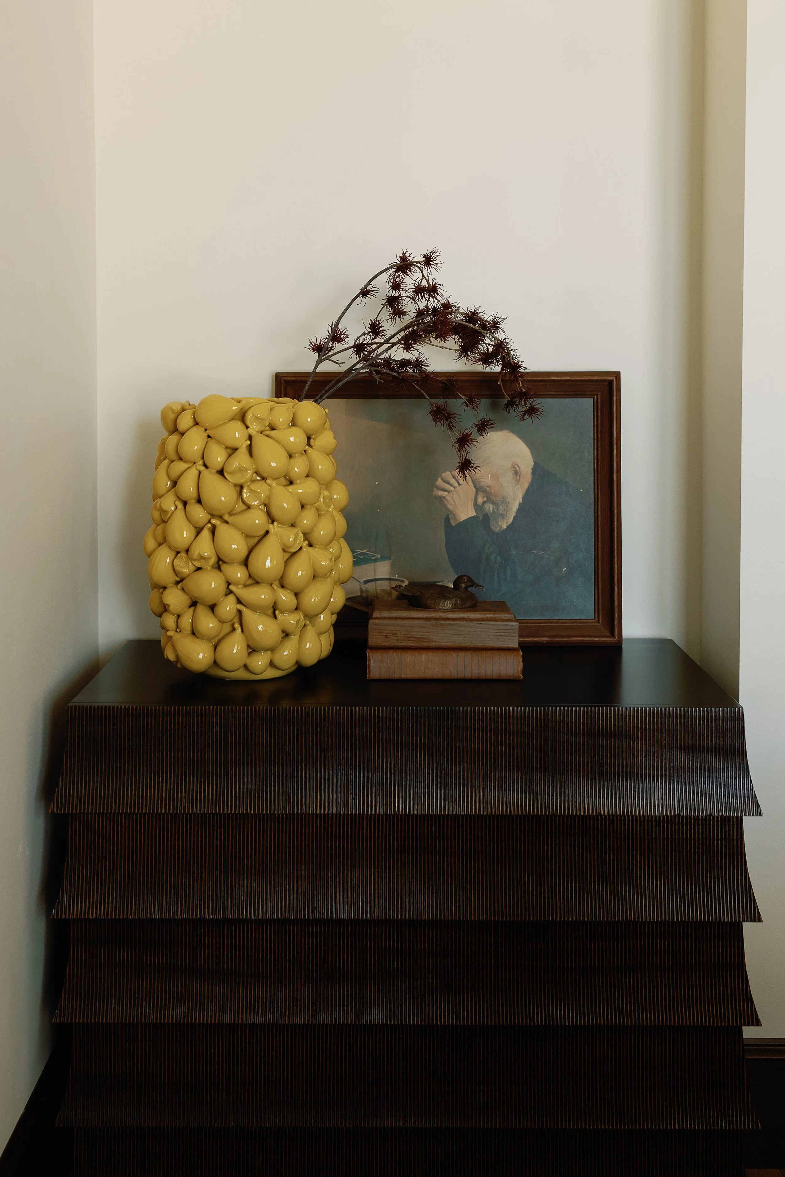 Decorative yellow vase with dried brown branches next to framed portrait of an elderly man resting his face on his hand, all on a dark wooden table against a white wall.
